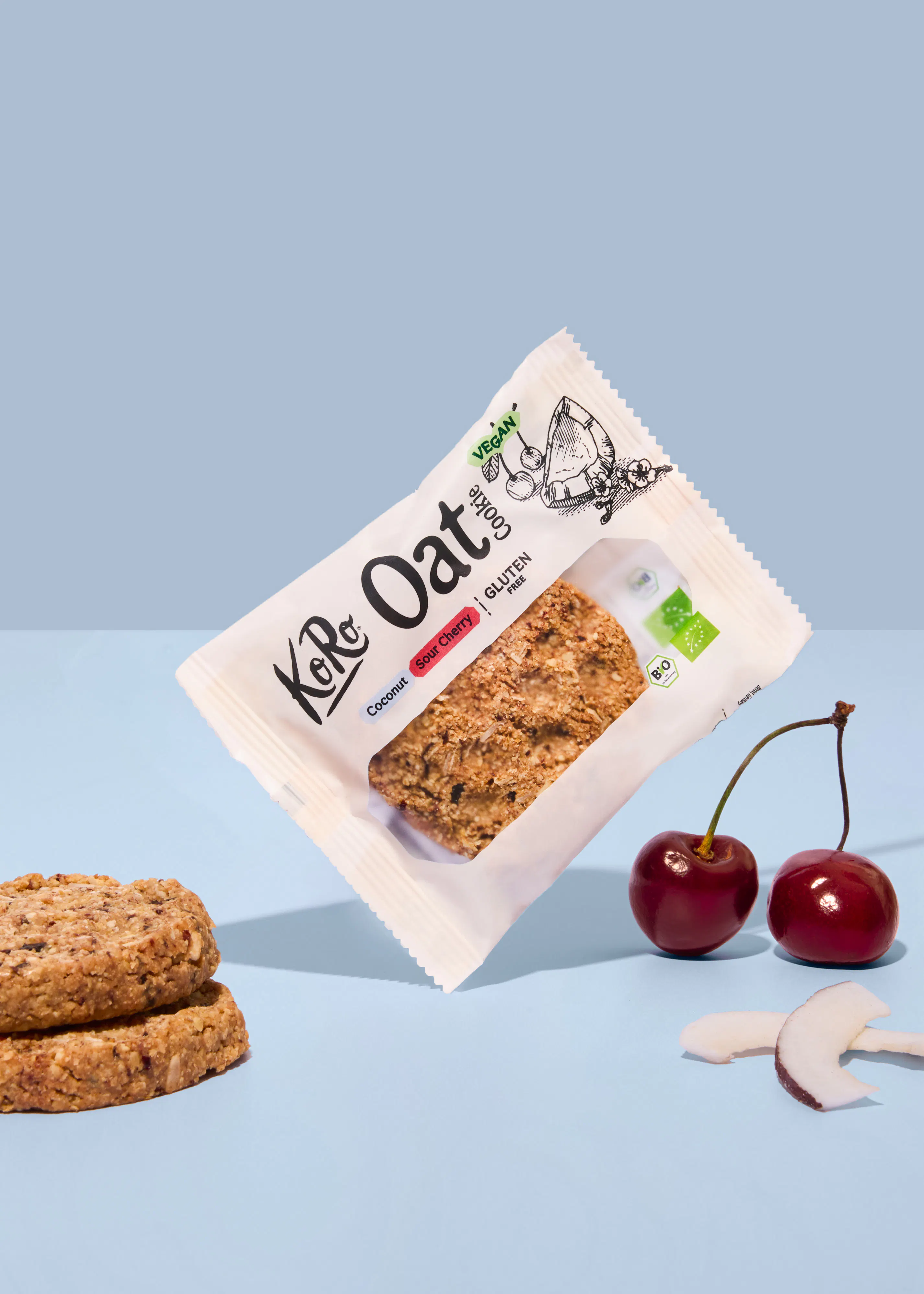 eine verpackung eines koro oat cookies mit kokosnuss und sauerkirsche neben zwei frischen kirschen und einem stück kokosnuss auf hellblauem hintergrund.