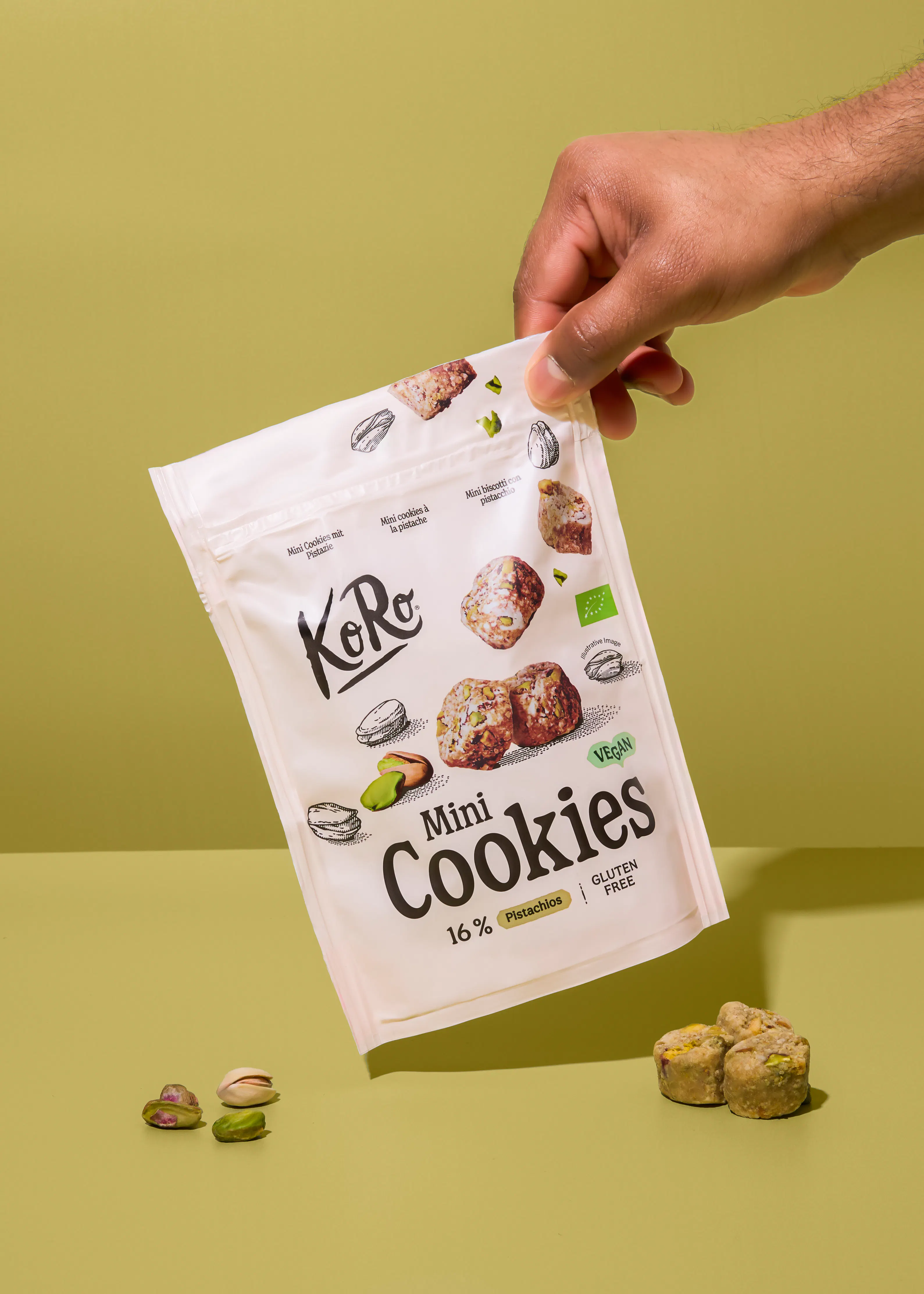 eine hand hält eine weiße tüte mit koro mini cookies mit pistazien über einem grünen hintergrund, daneben liegen lose pistazien und kekse.
