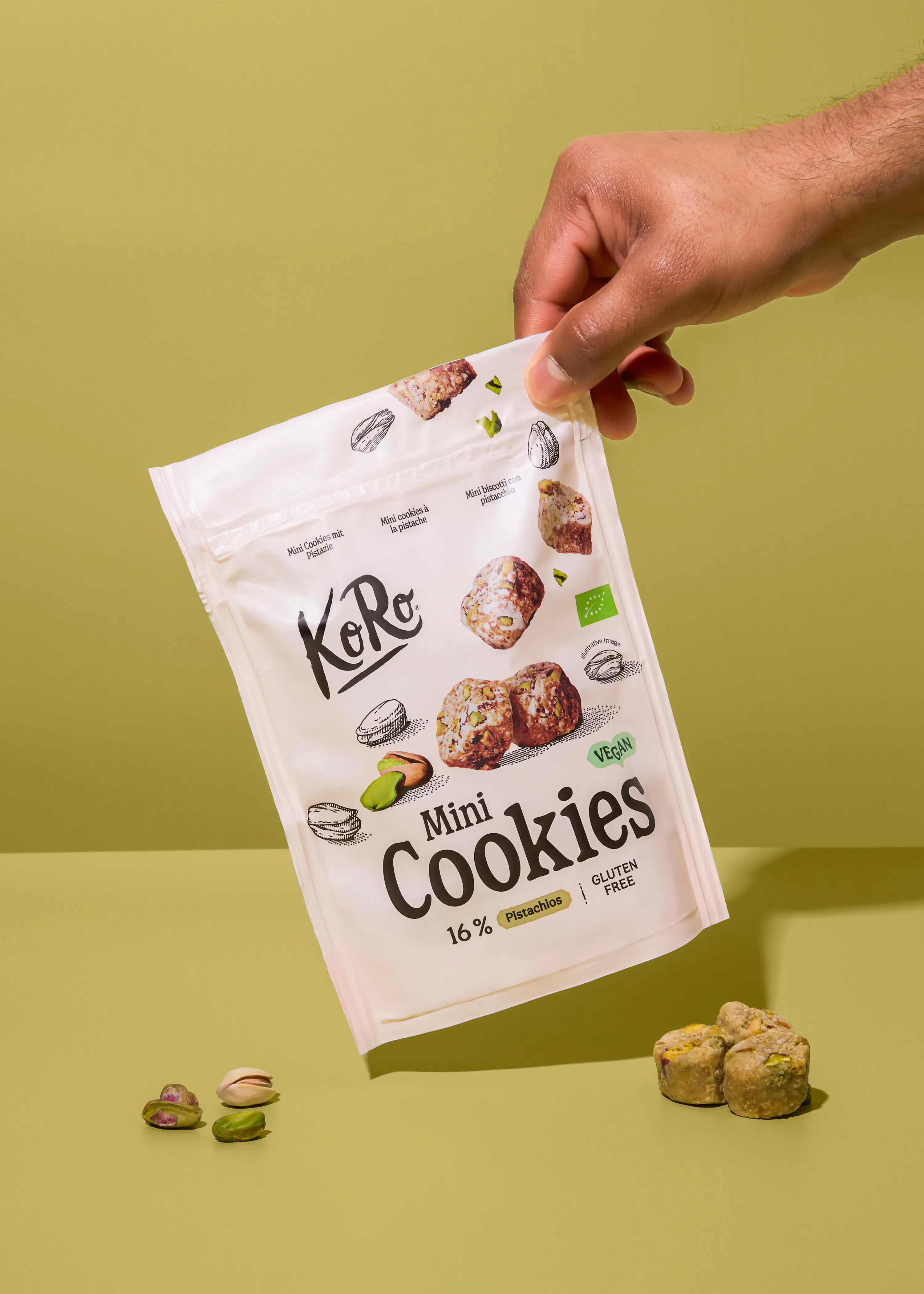 une main tient un sachet blanc de mini cookies ko ro aux pistaches, avec des pistaches et des cookies éparpillés sur une surface verte.