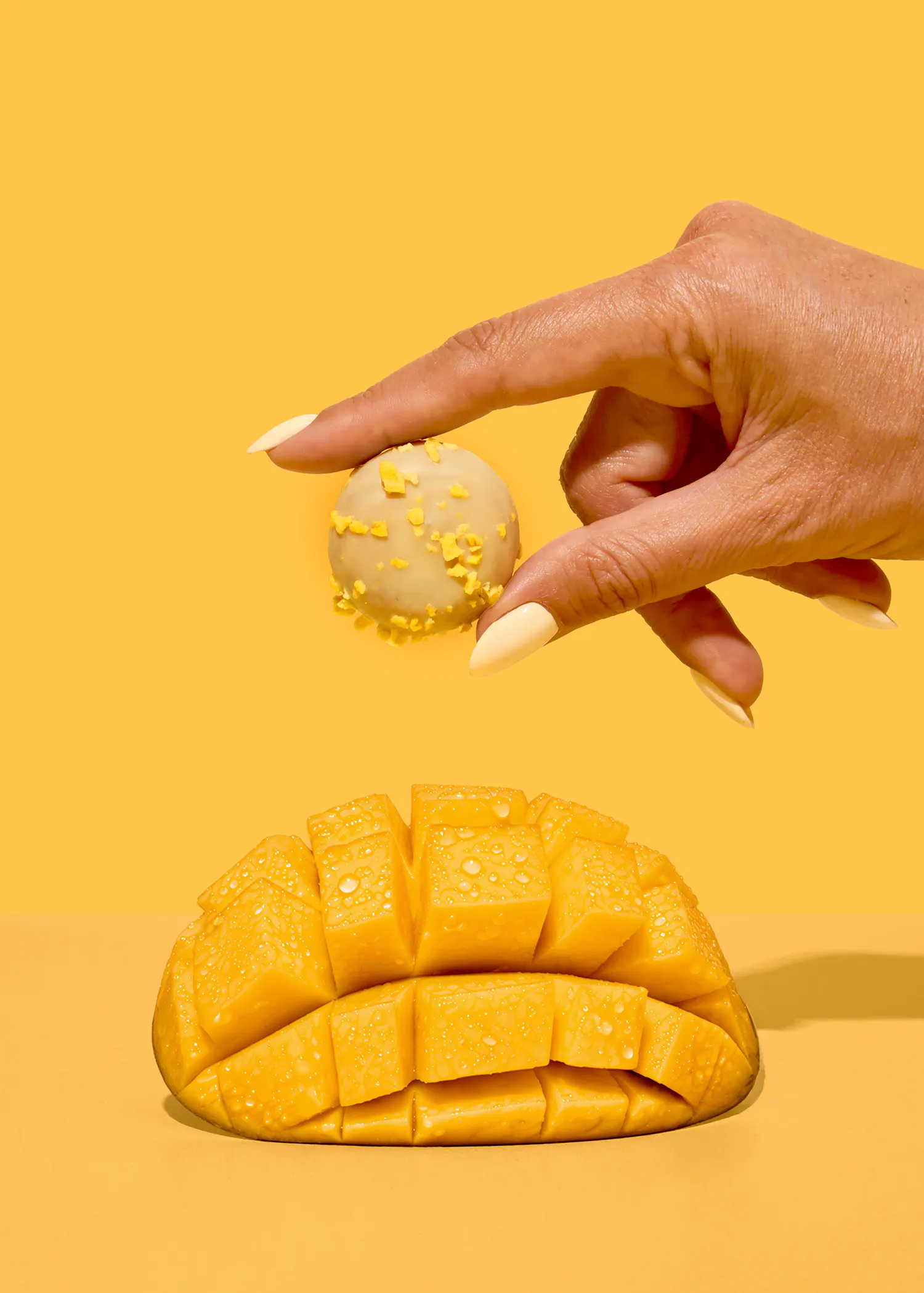 eine hand mit hellen nägeln hält einen hellen trüffel mit gelben streuseln über einer gewürfelten mango mit wassertropfen auf einem gelben hintergrund.