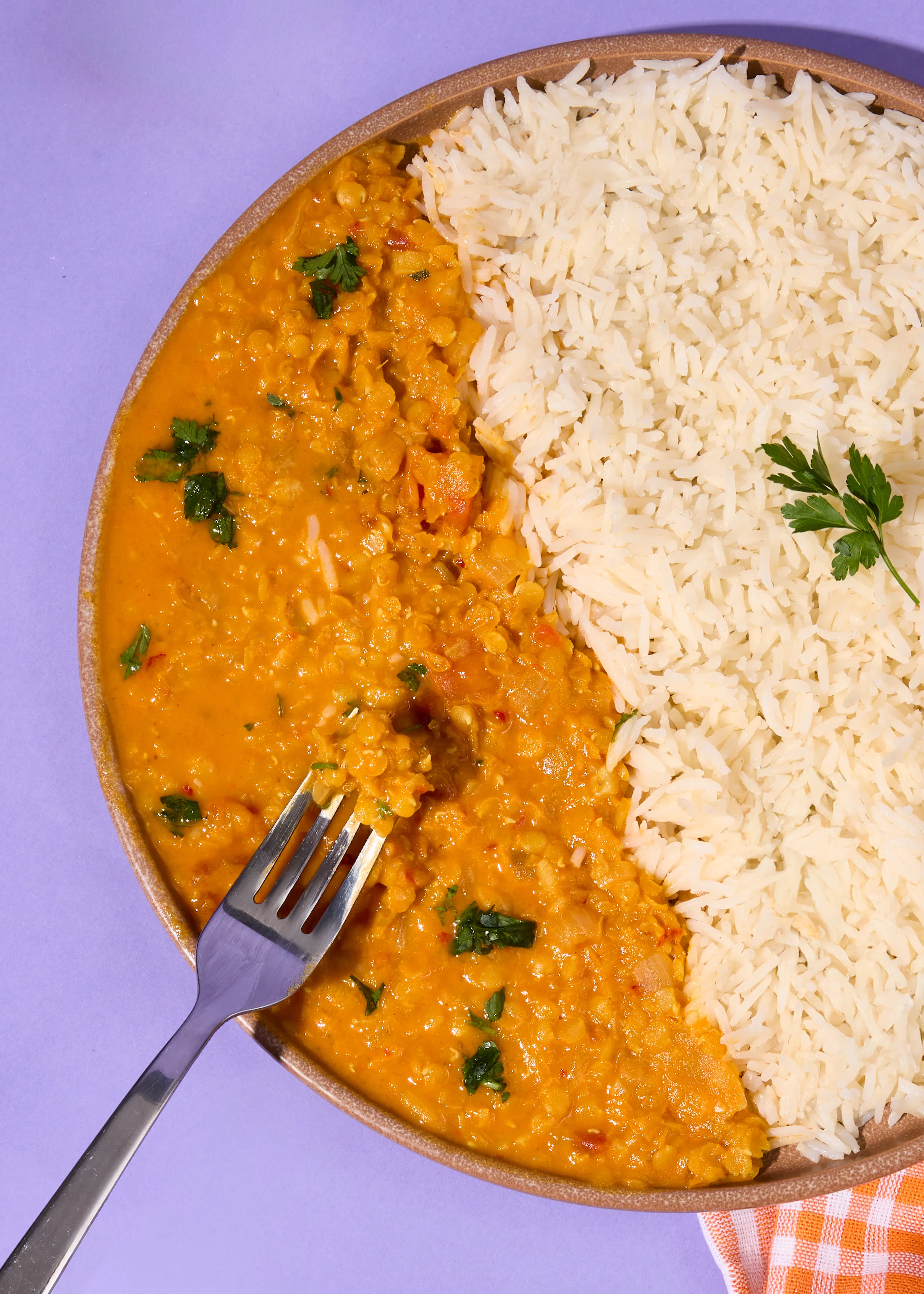 ein teller mit orangefarbenem linsencurry, garniert mit frischen kräutern, und weißem reis auf einem lila hintergrund, wobei eine gabel in das curry taucht.