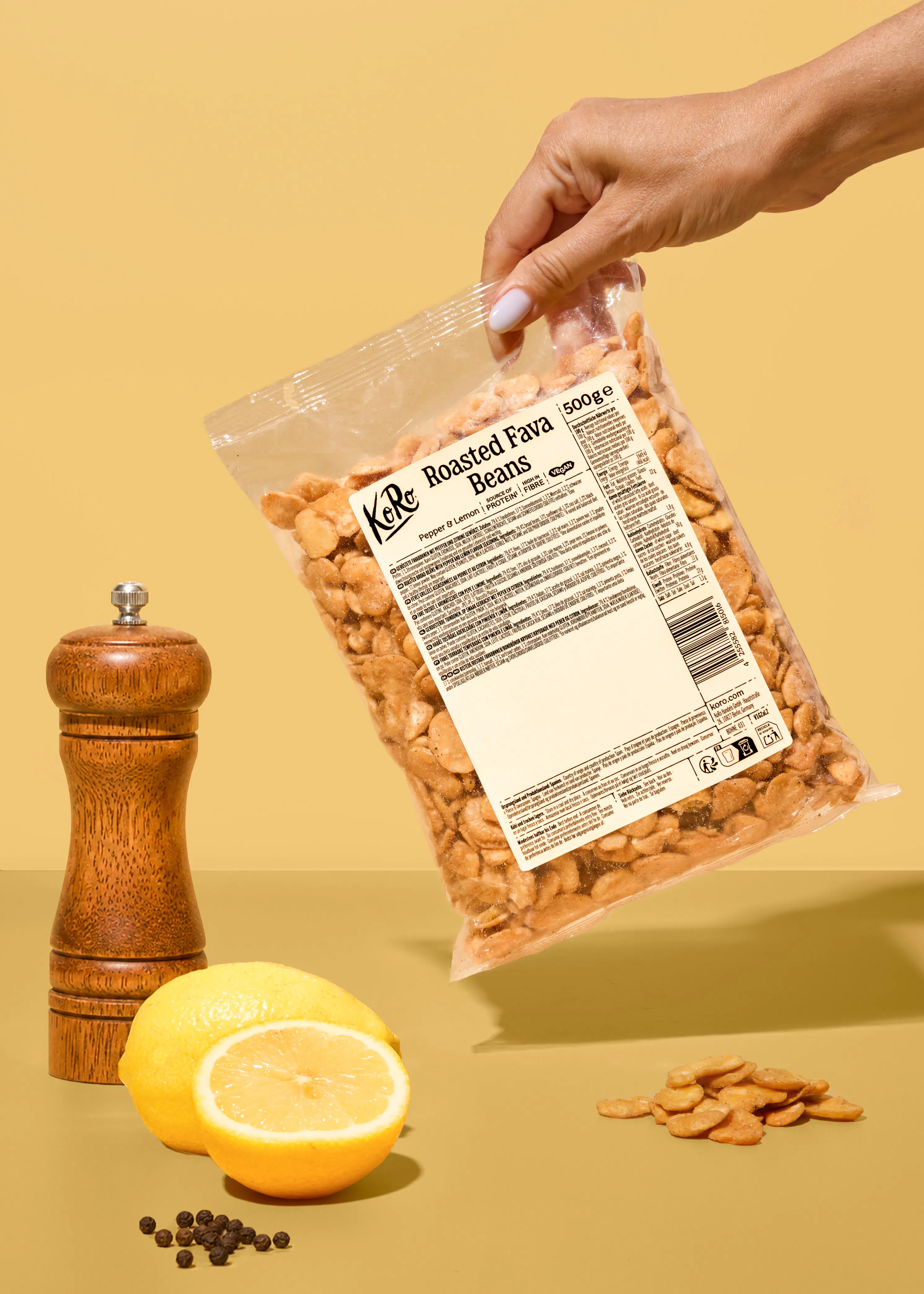 une main tient un sachet de fèves grillées au poivre et au citron de la marque koro au-dessus d'un moulin à poivre et de citrons frais sur un fond beige.