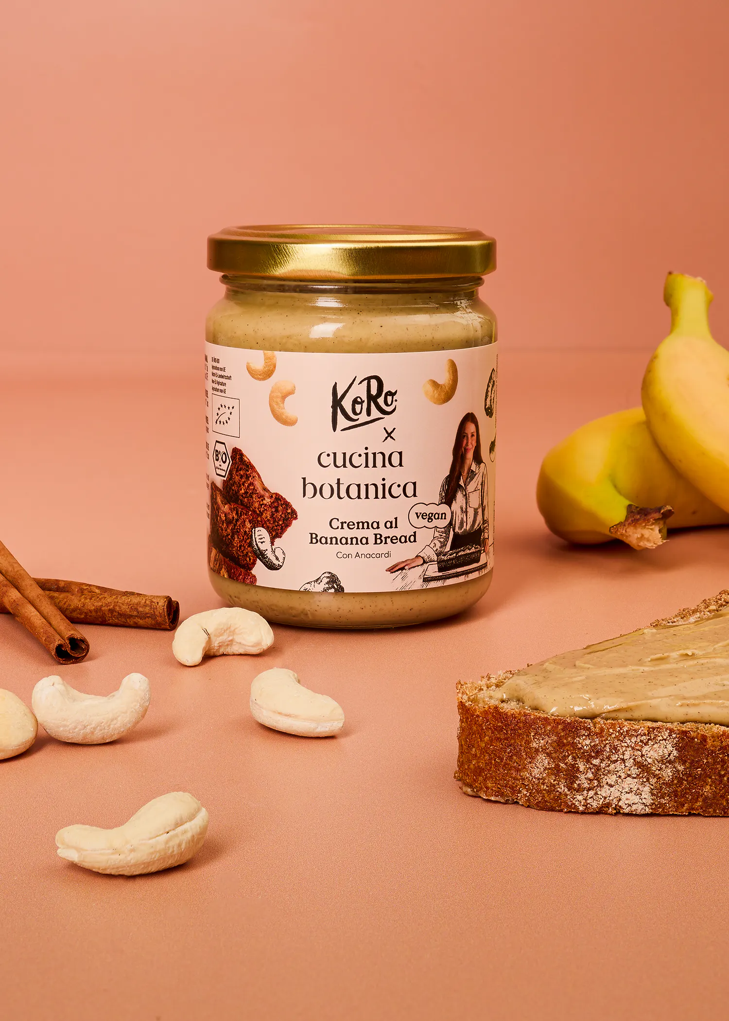 ein glas koro x cucina botanica banana bread creme umgeben von bananen, cashews und zimtstangen auf einem rosa hintergrund.