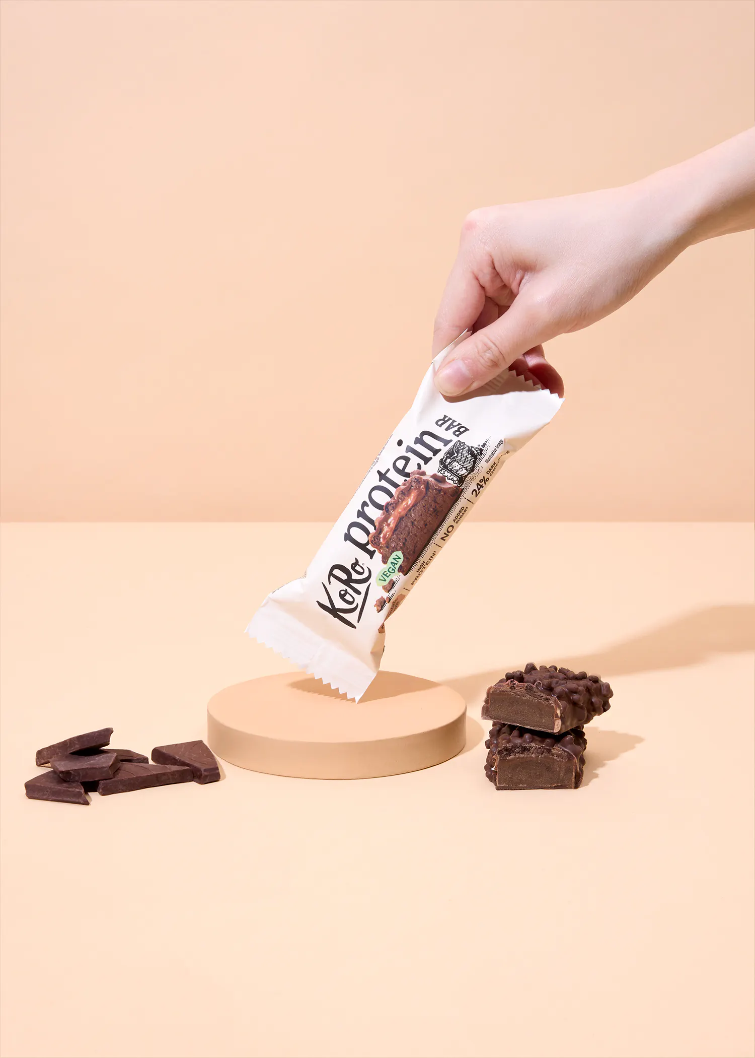 eine hand hält einen weißen proteinriegel über einem beigen podest, daneben liegen schokoladenstücke und ein aufgeschnittener riegel auf einem hellen hintergrund.