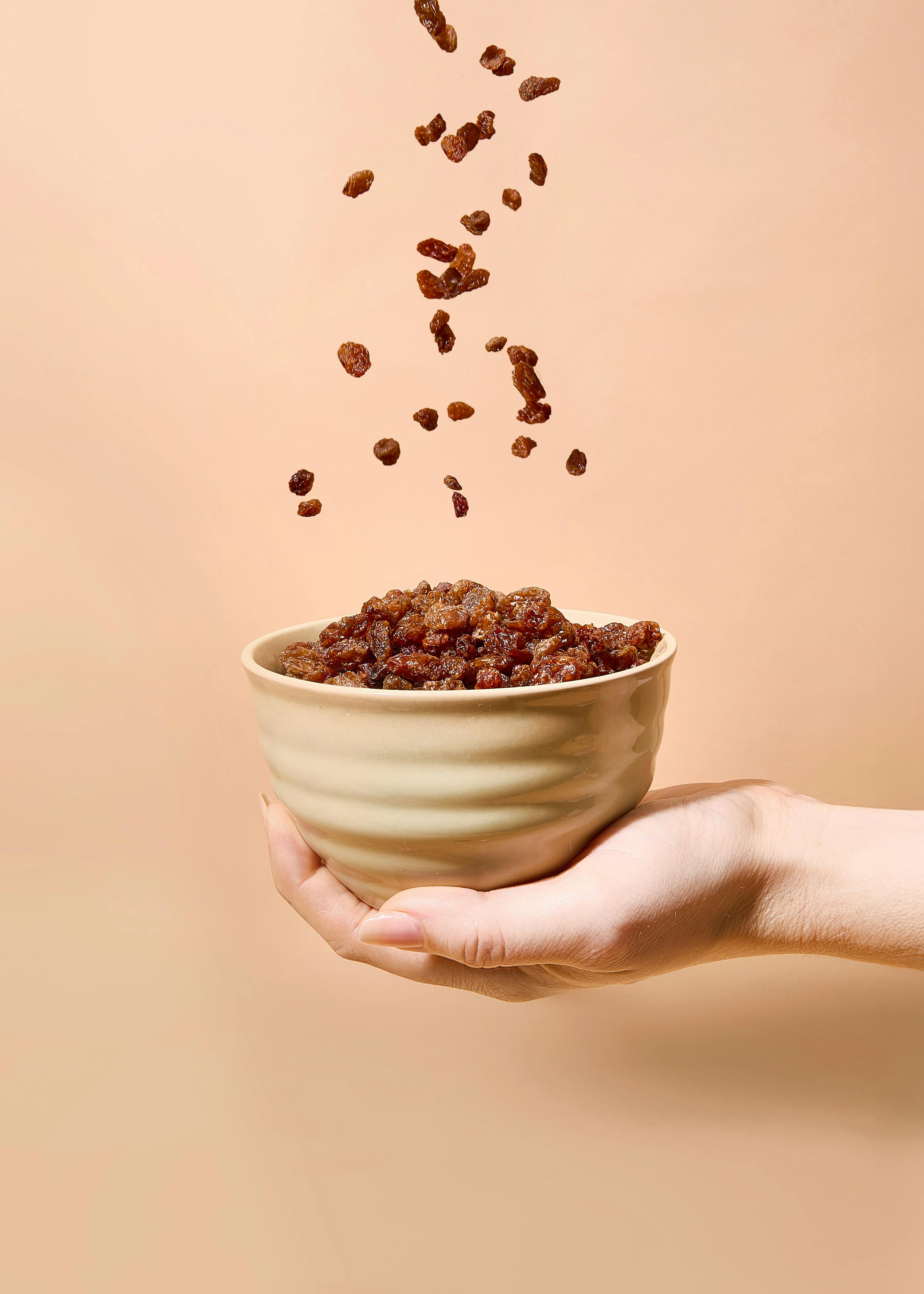 une main tenant un bol rempli de raisins secs, avec d'autres raisins secs tombant dans le bol.