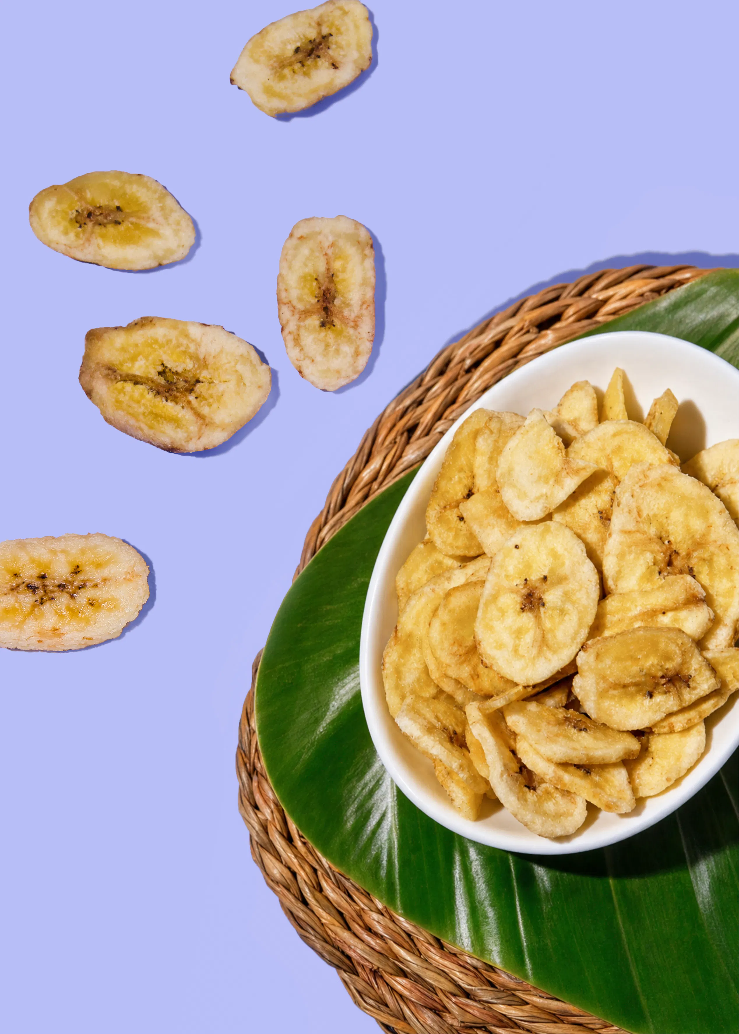 bananenchips in einer weißen schale auf einem bananenblatt und einer geflochtenen matte, mit einigen chips auf einem lila hintergrund verstreut.