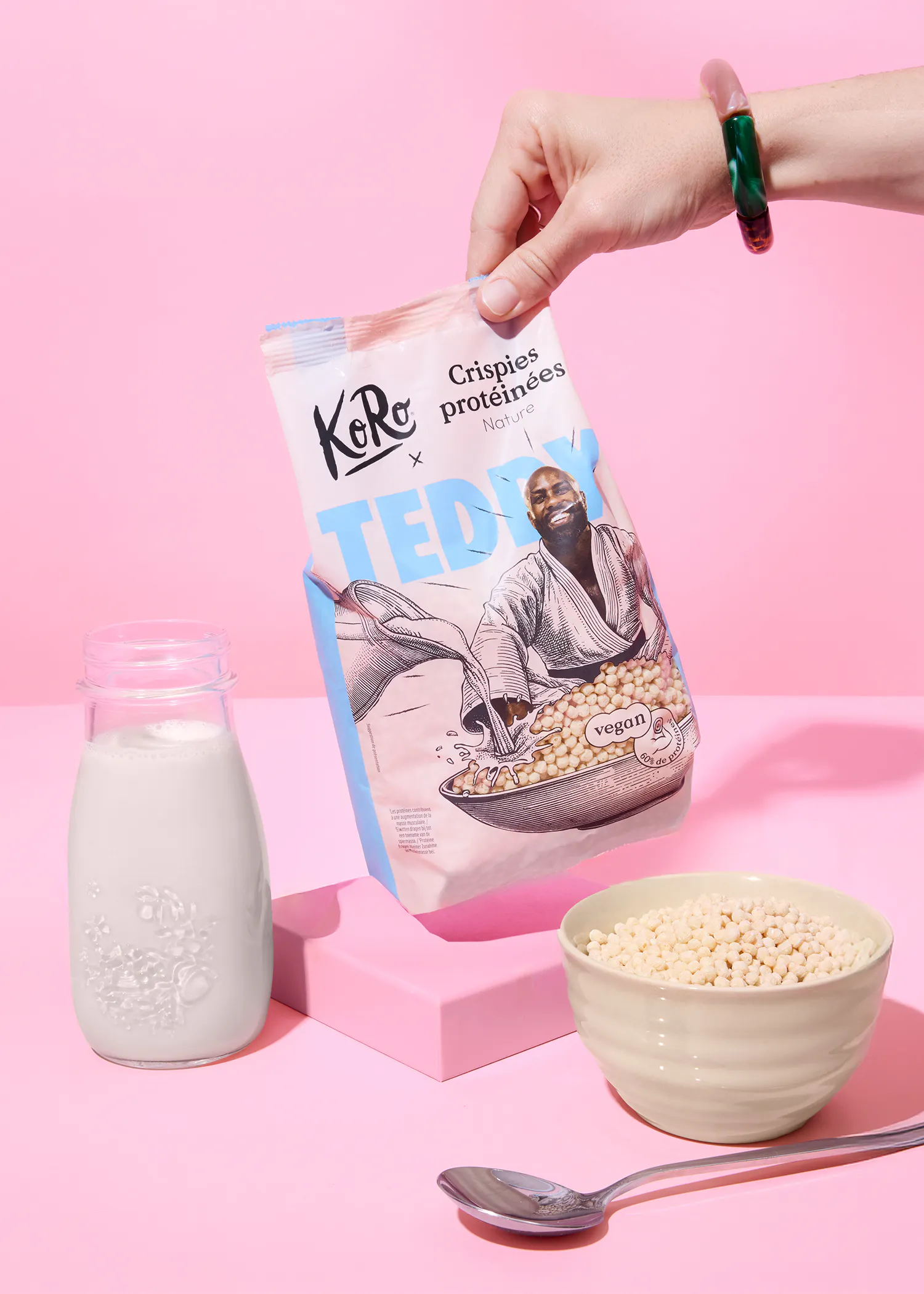 une main tient un sac de crispies protéinées koro x teddy, versant le contenu dans un bol, avec une bouteille de lait et une cuillère sur un fond rose.