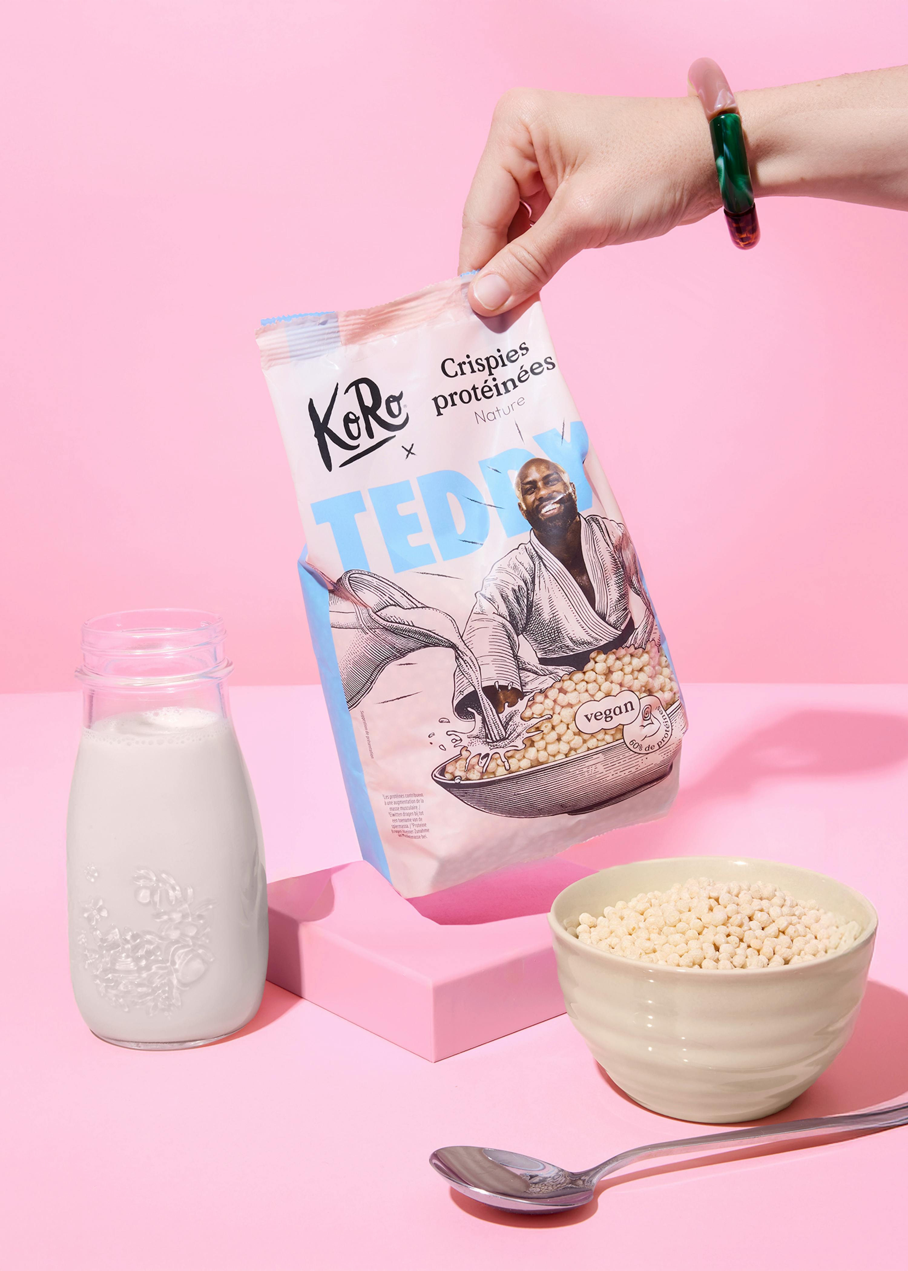 une main tient un sac de crispies protéinées koro x teddy, versant le contenu dans un bol, avec une bouteille de lait et une cuillère sur un fond rose.