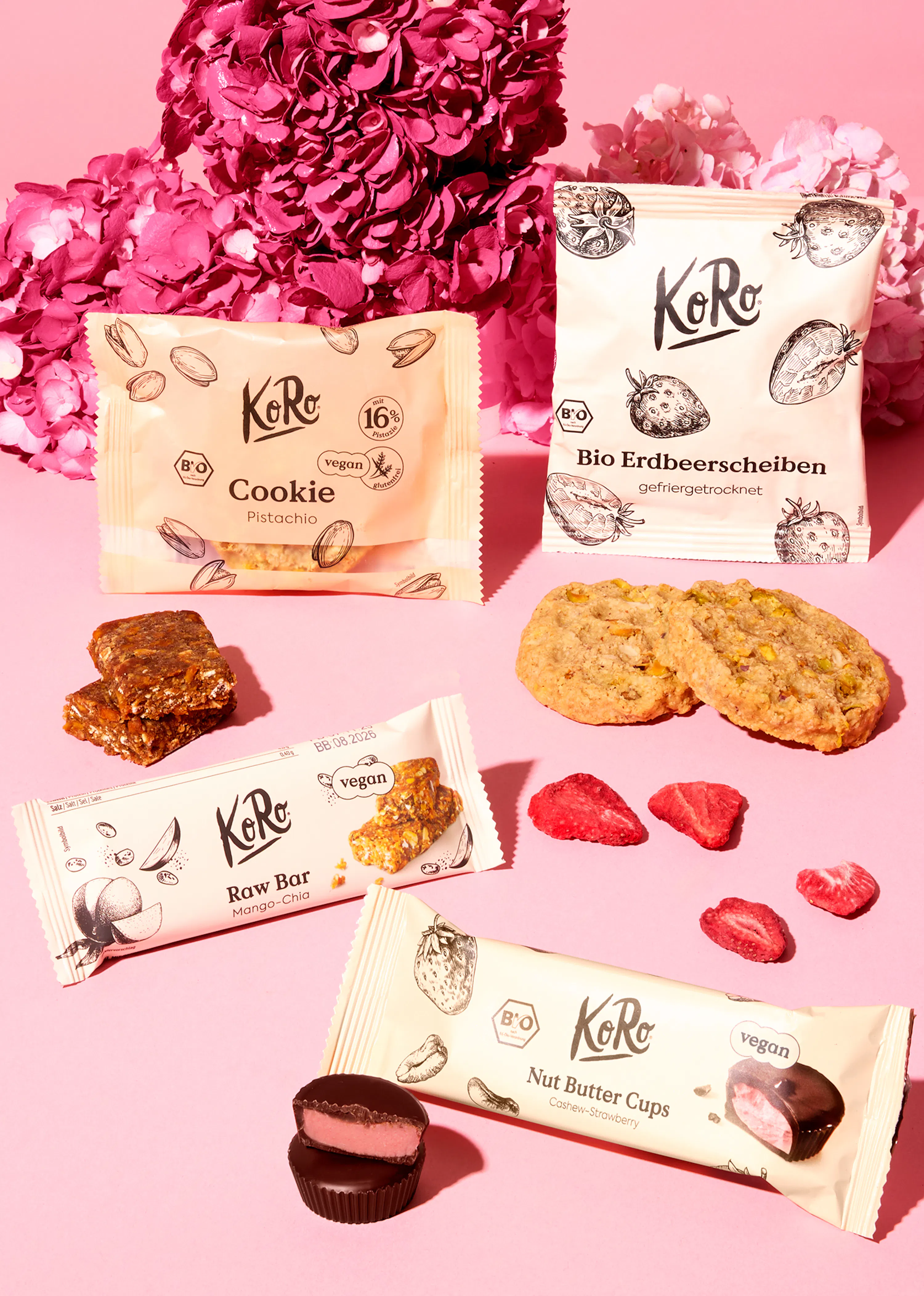 verschiedene vegane koro-snacks, darunter pistazienkekse, gefriergetrocknete erdbeeren, mango-chia-riegel und cashew-erdbeer-nussbutter-cups, sind auf einem pinken hintergrund mit pinken blumen arrangiert.
