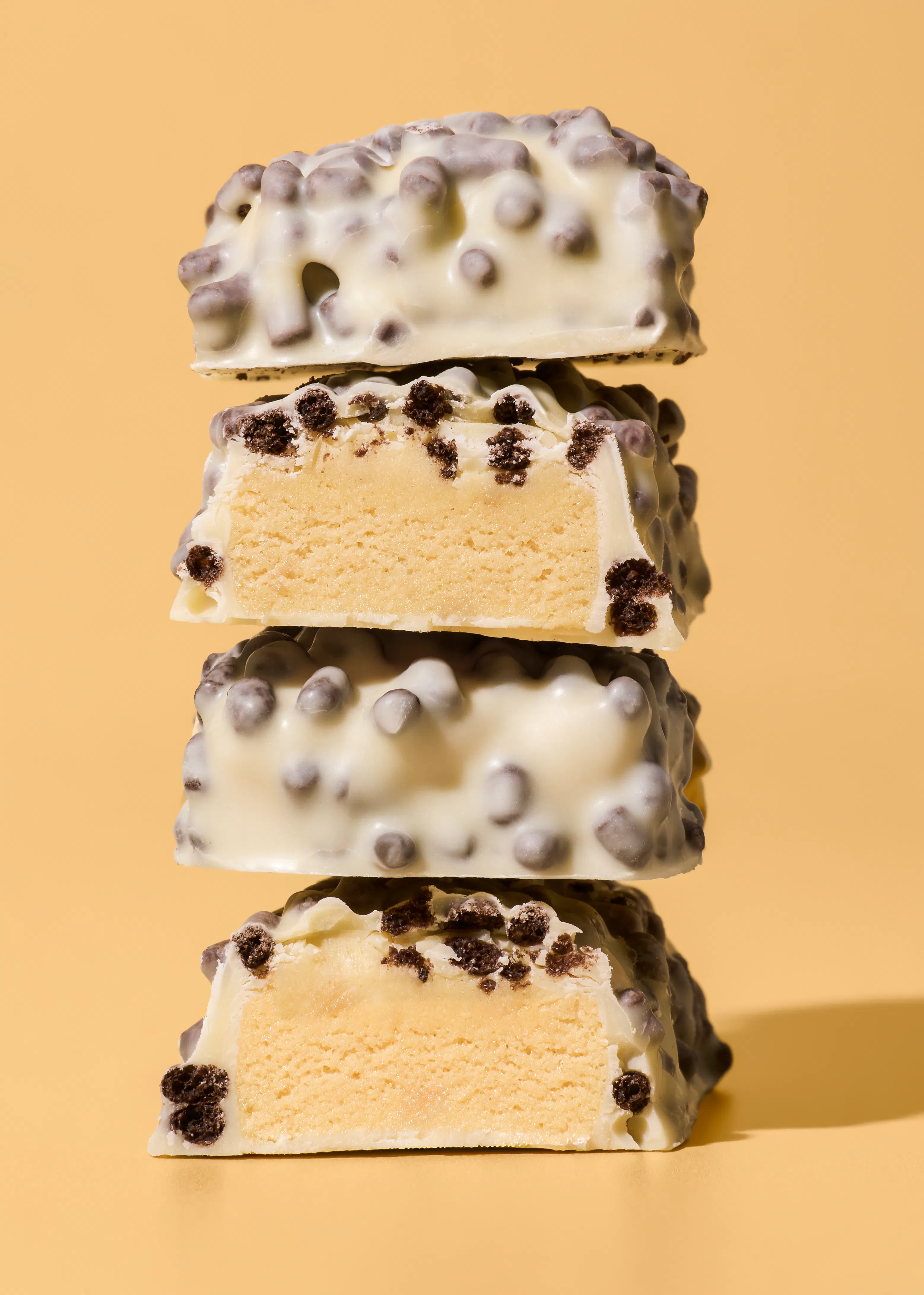 a stack of four white chocolate-covered bars with dark cookie crumbles, two of which are cut in half to reveal a light-colored interior, against a yellow background.