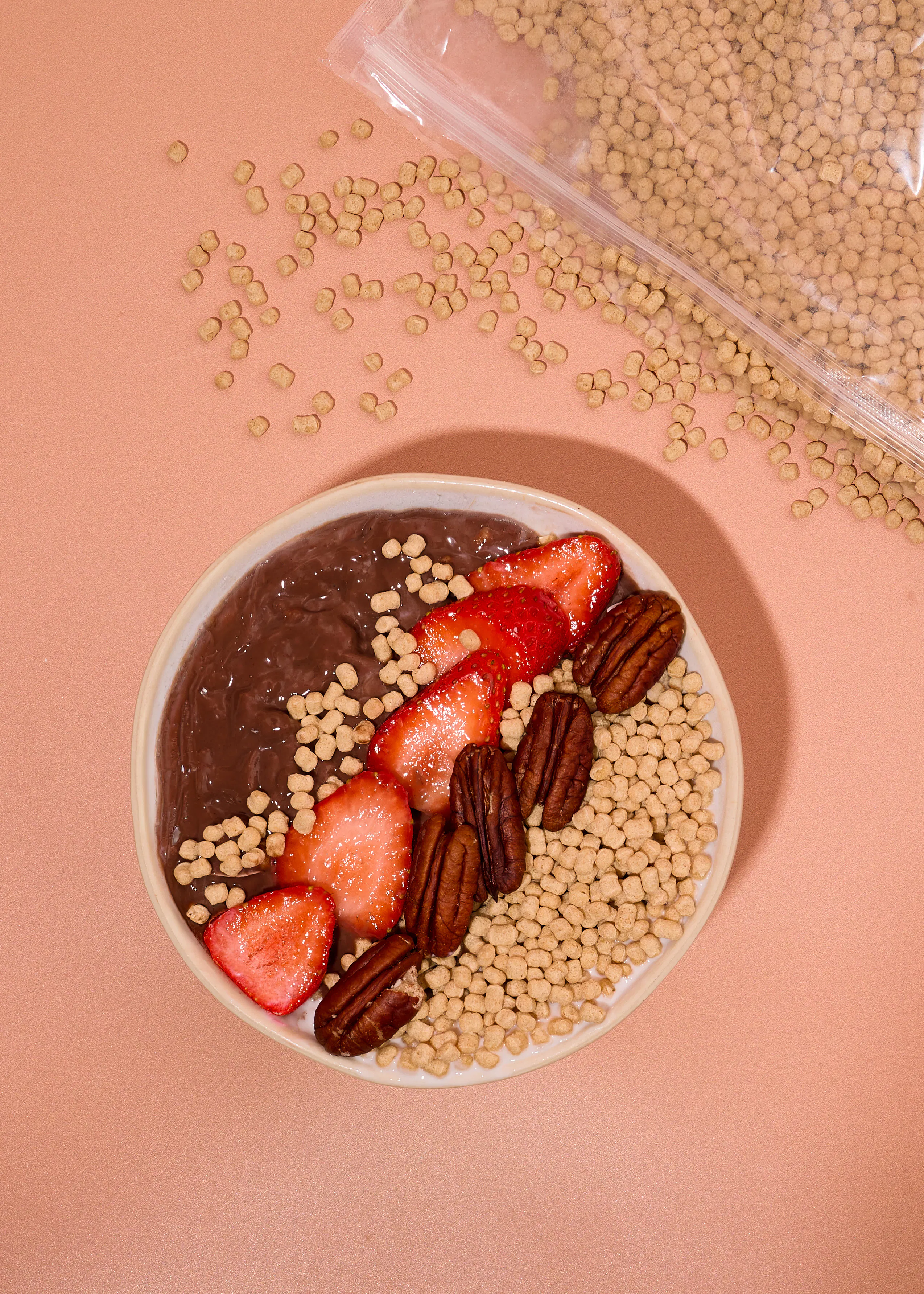 una ciotola di crema al cioccolato guarnita con fragole a fette, noci pecan e cereali croccanti su uno sfondo color pesca con una busta di cereali aperta accanto.
