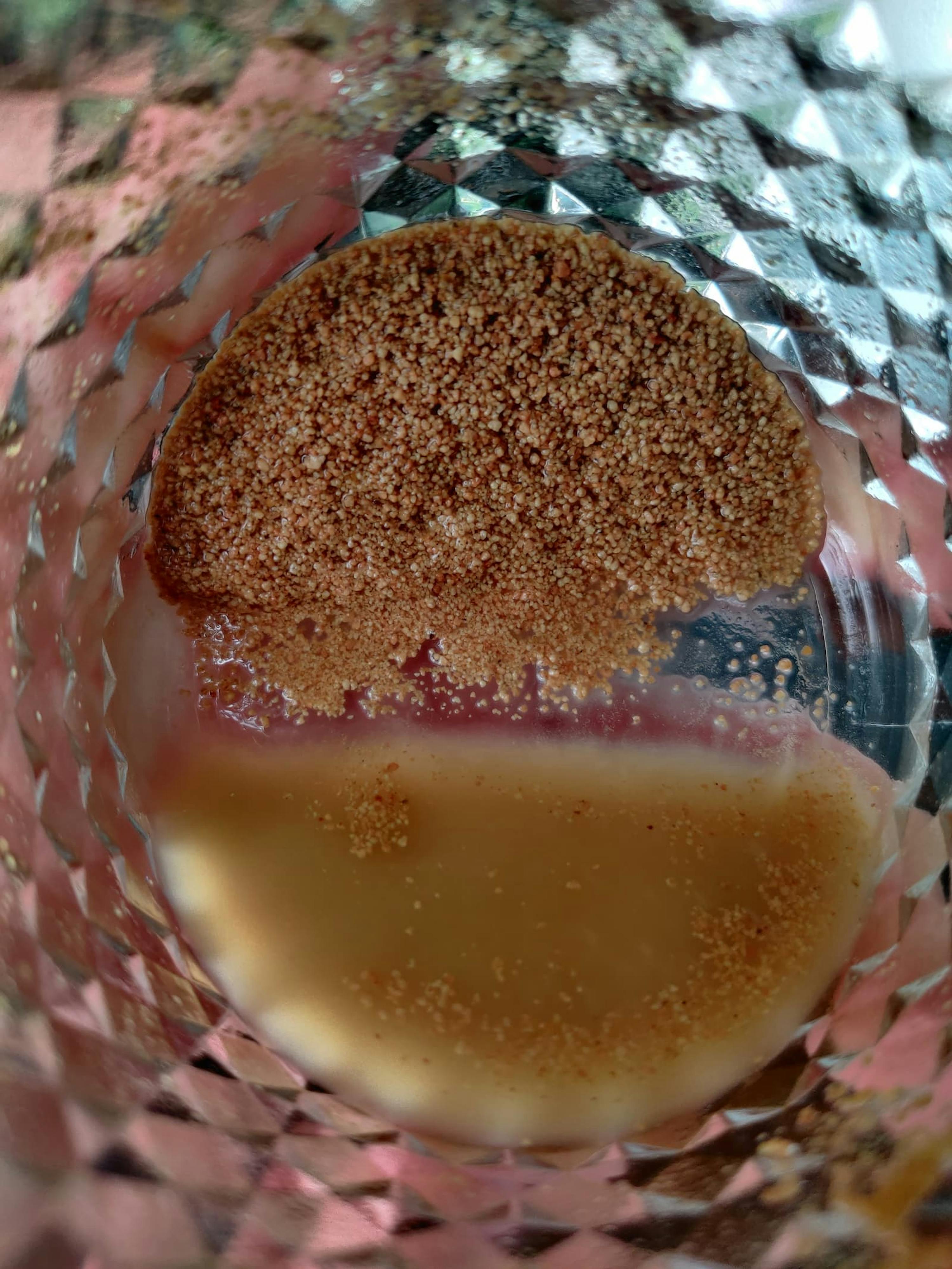 gros plan sur une couche de poudre granuleuse brune flottant sur un liquide jaunâtre à l'intérieur d'un verre texturé.