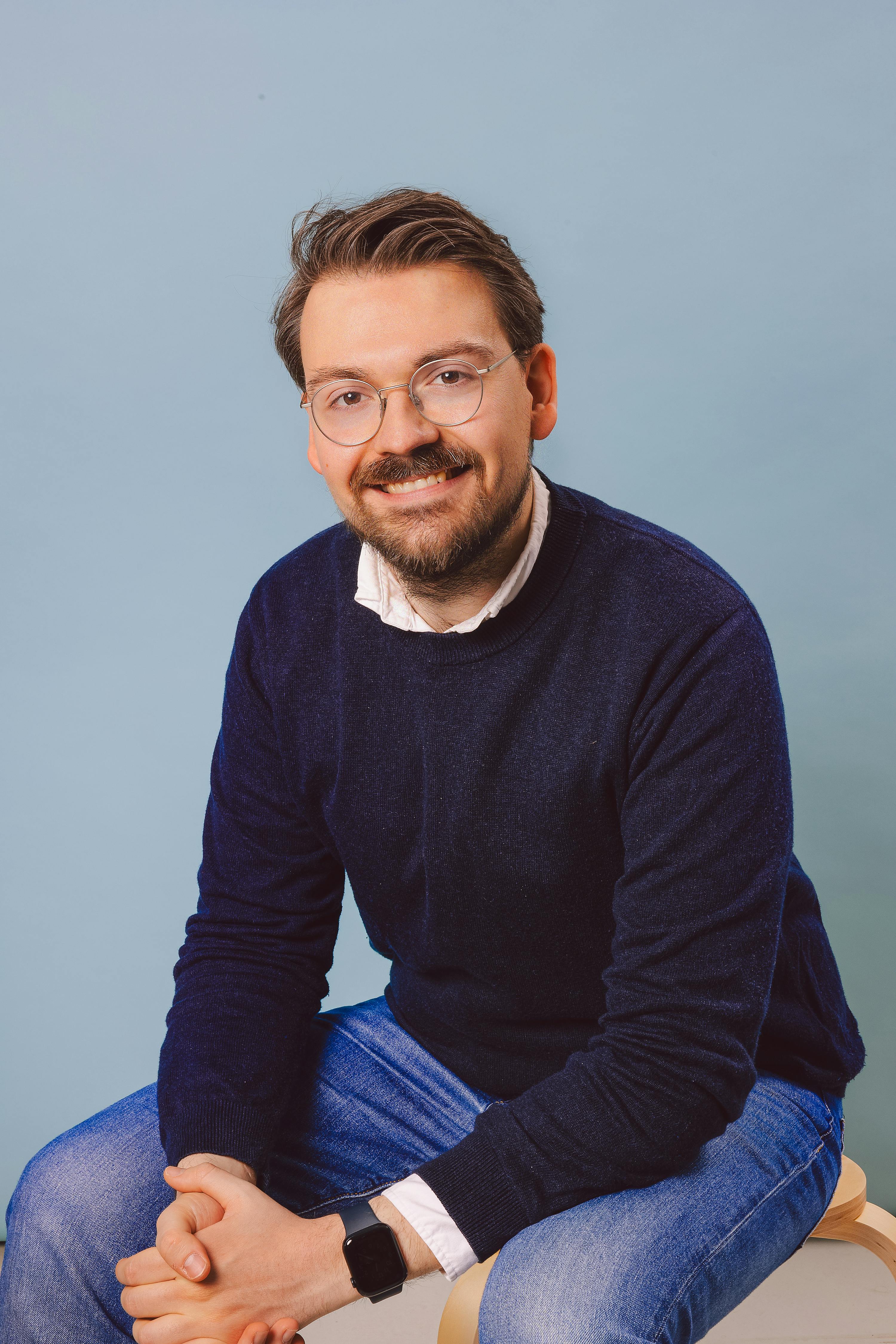 un homme portant un pull bleu et des lunettes est assis avec ses mains croisées