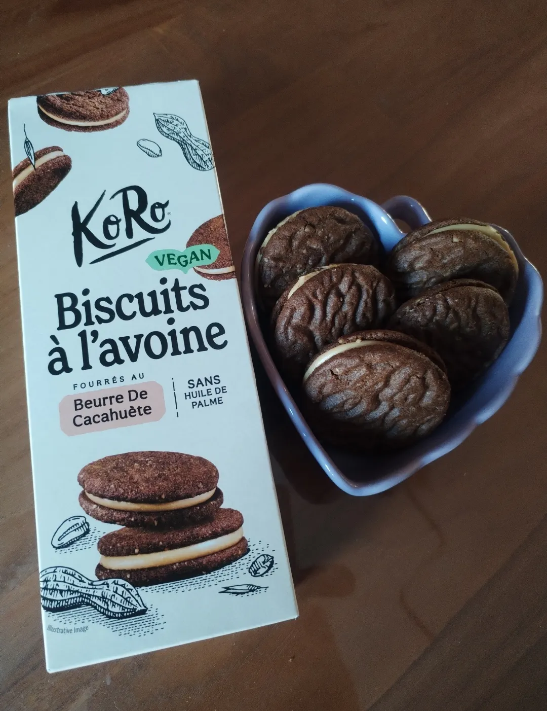 una confezione di biscotti vegani koro all'avena ripieni di burro di arachidi accanto a una ciotola a forma di cuore contenente alcuni biscotti.
