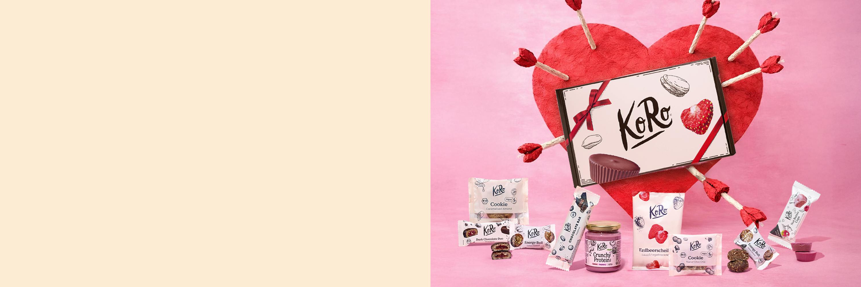 ein rotes herz mit pfeilen und einer koro-karte, umgeben von verschiedenen koro-produkten wie keksen, riegeln, aufstrich und nussbutter-cups, auf einem rosa hintergrund.
