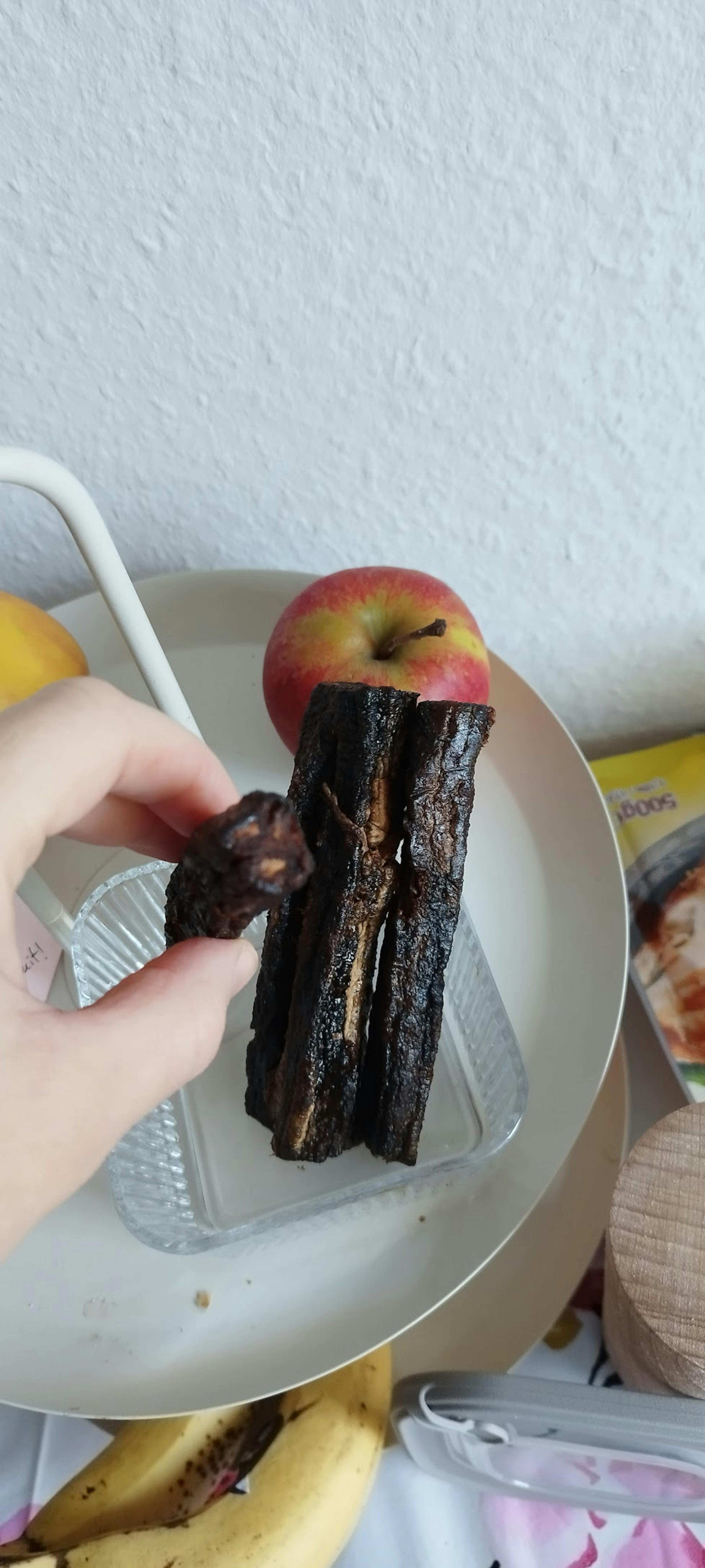 a hand holds a dark, dried fruit, with additional dried fruit, an apple, and a banana on a white tray.