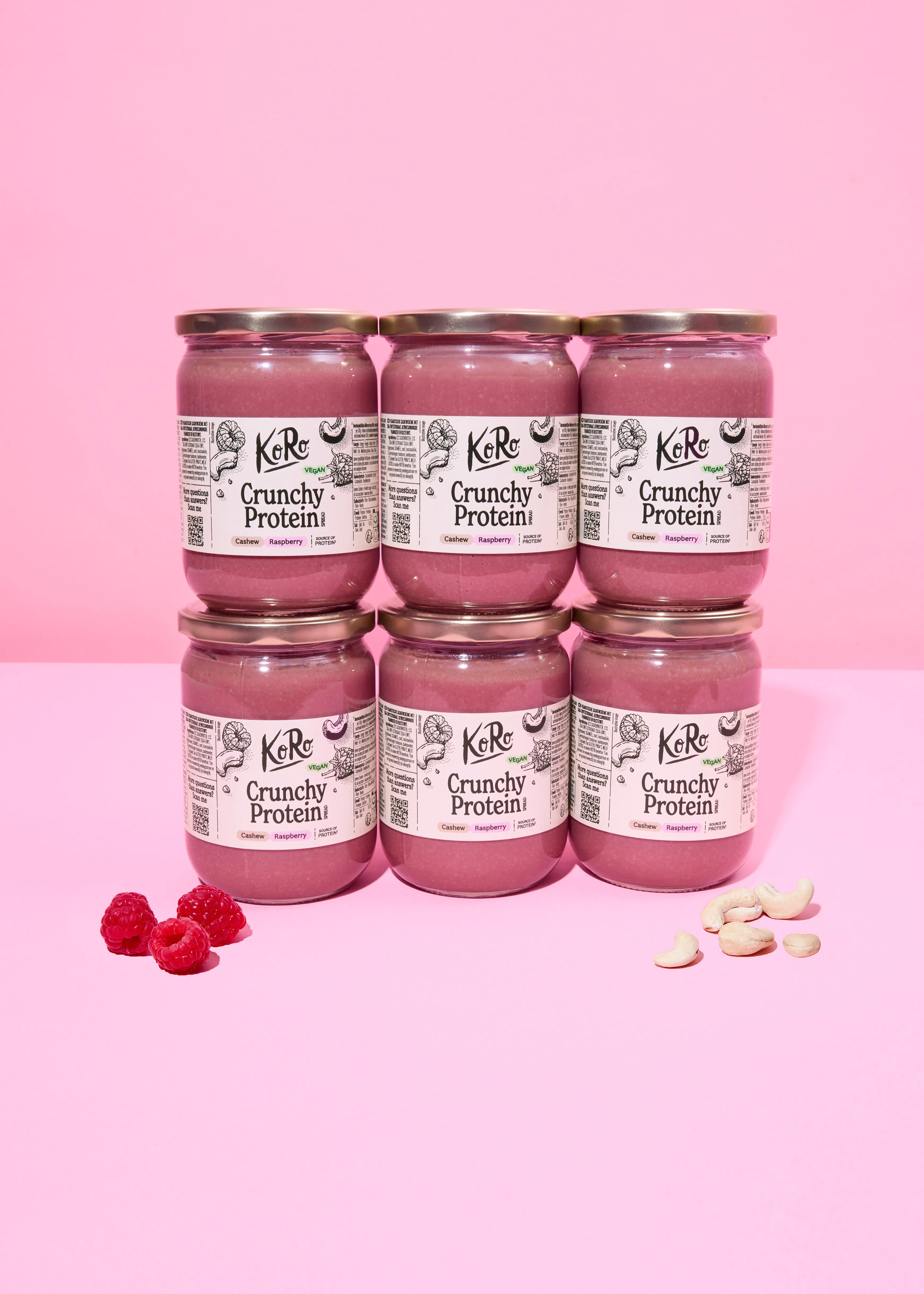 sechs gläser koro crunchy protein cashew-himbeer-aufstrich, in zwei reihen gestapelt auf einem rosa hintergrund, mit frischen himbeeren und cashewnüssen.