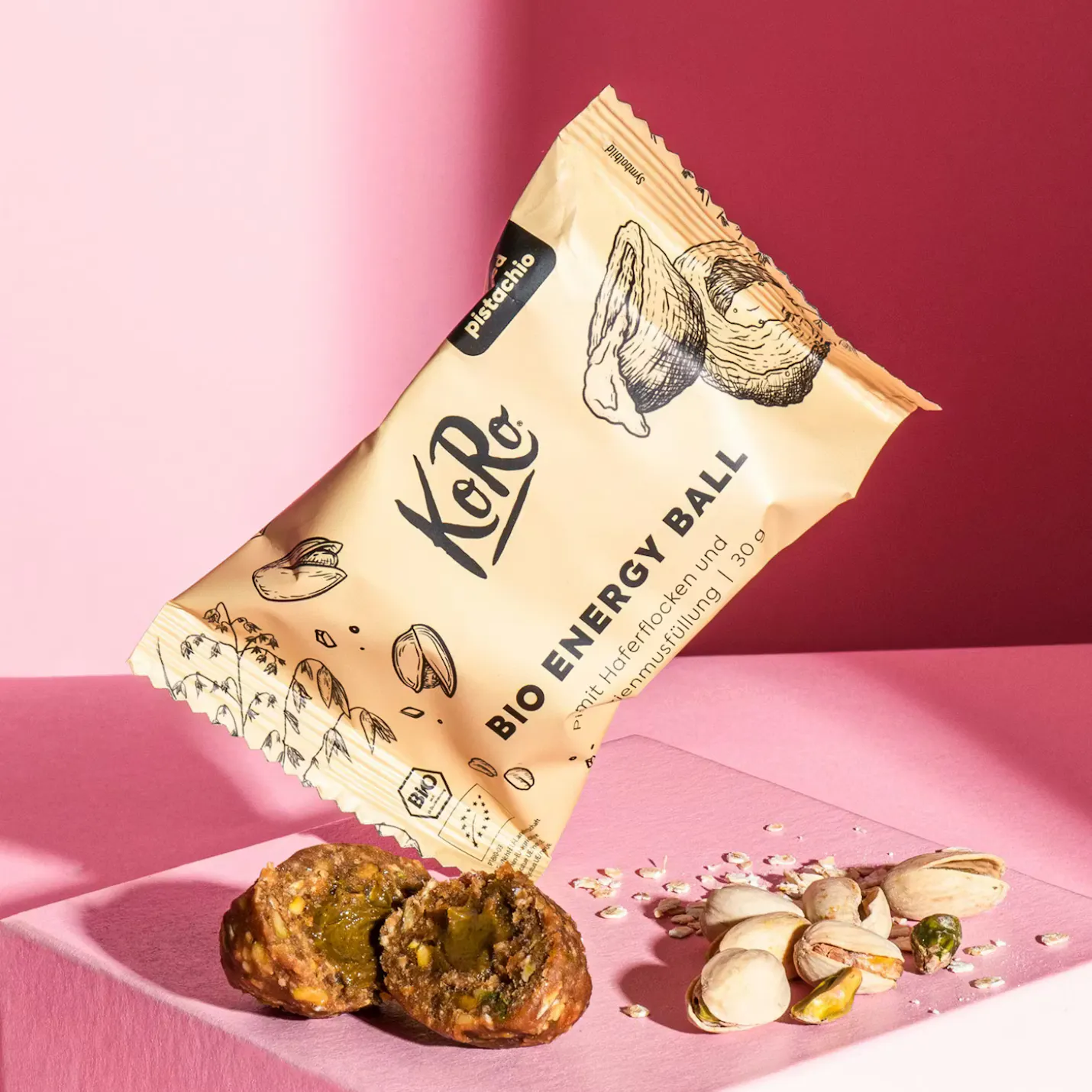un paquete de bola energética de pistacho koro bio flotando sobre una superficie rosa con dos bolas energéticas cortadas, pistachos y avena esparcidos.