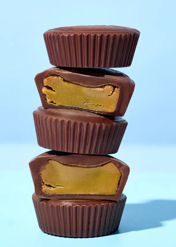 une pile de quatre coupes au beurre de cacahuète enrobées de chocolat, dont deux sont coupées en deux pour révéler la garniture crémeuse, sur un fond bleu clair.