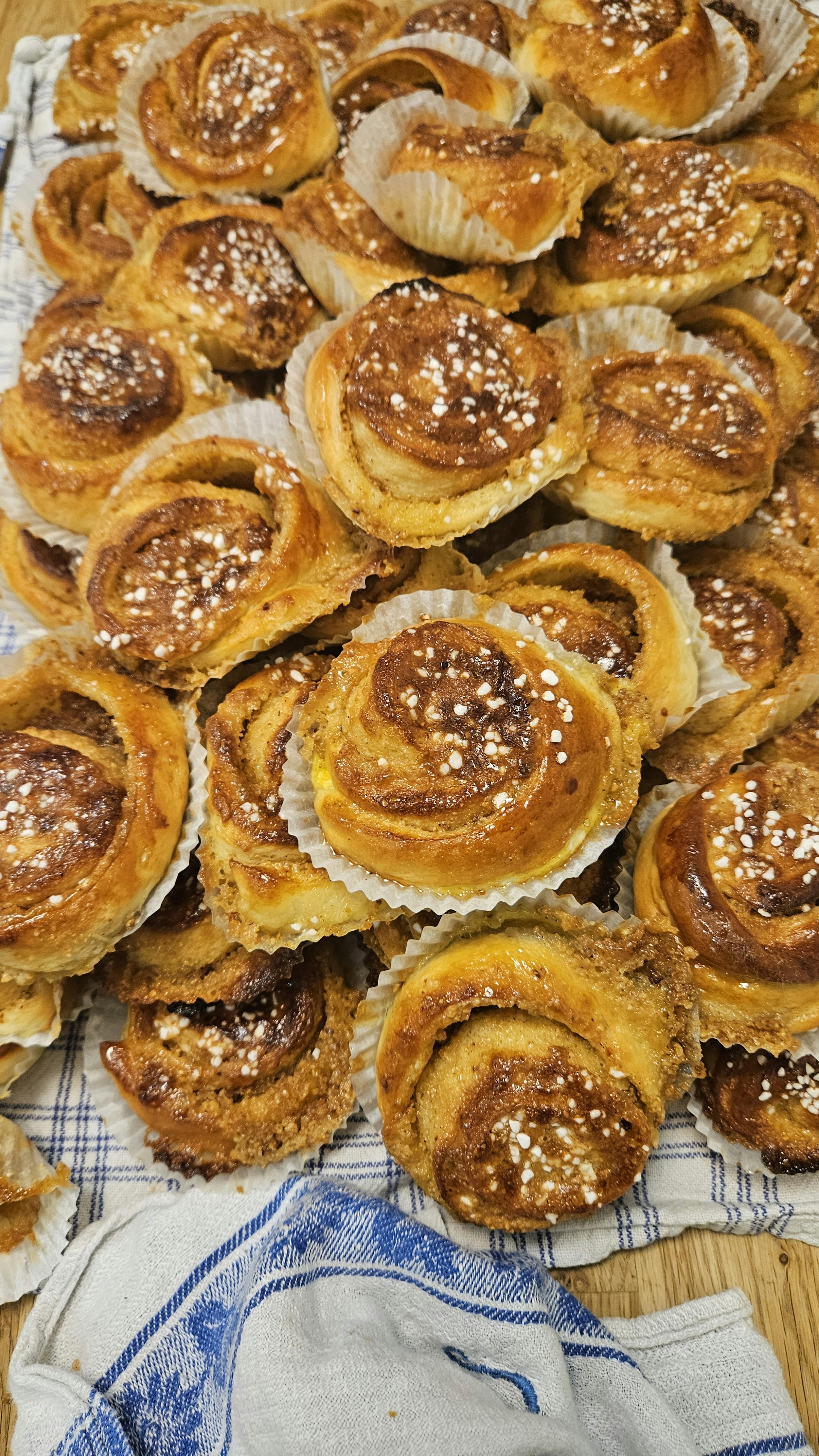 viele goldbraune zimtschnecken mit hagelzucker, einige in papierförmchen, liegen auf einem tisch neben einem blau-weiß karierten tuch.
