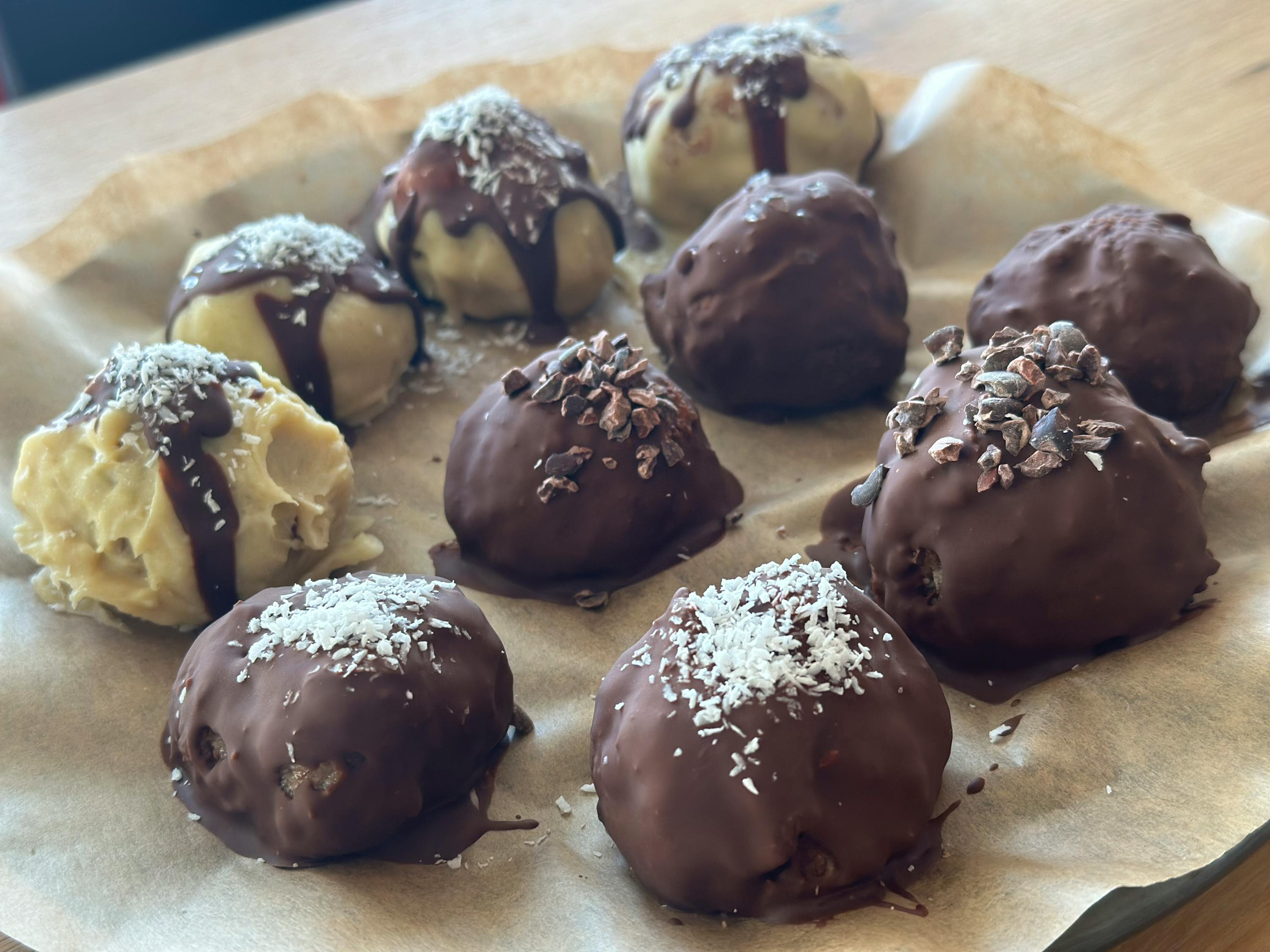 mehrere runde pralinen auf backpapier, einige hell mit schokoladenüberzug und kokosraspeln, andere dunkel mit schokolade und kakaonibs oder kokosraspeln.