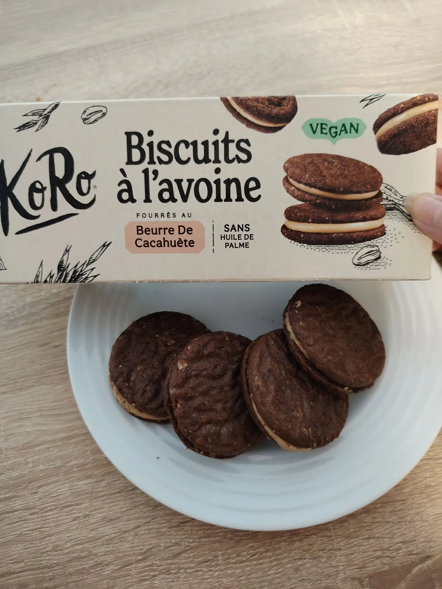 una mano tiene una confezione di biscotti vegani koro all'avena ripieni di burro di arachidi sopra un piattino con alcuni biscotti disposti a semicerchio.