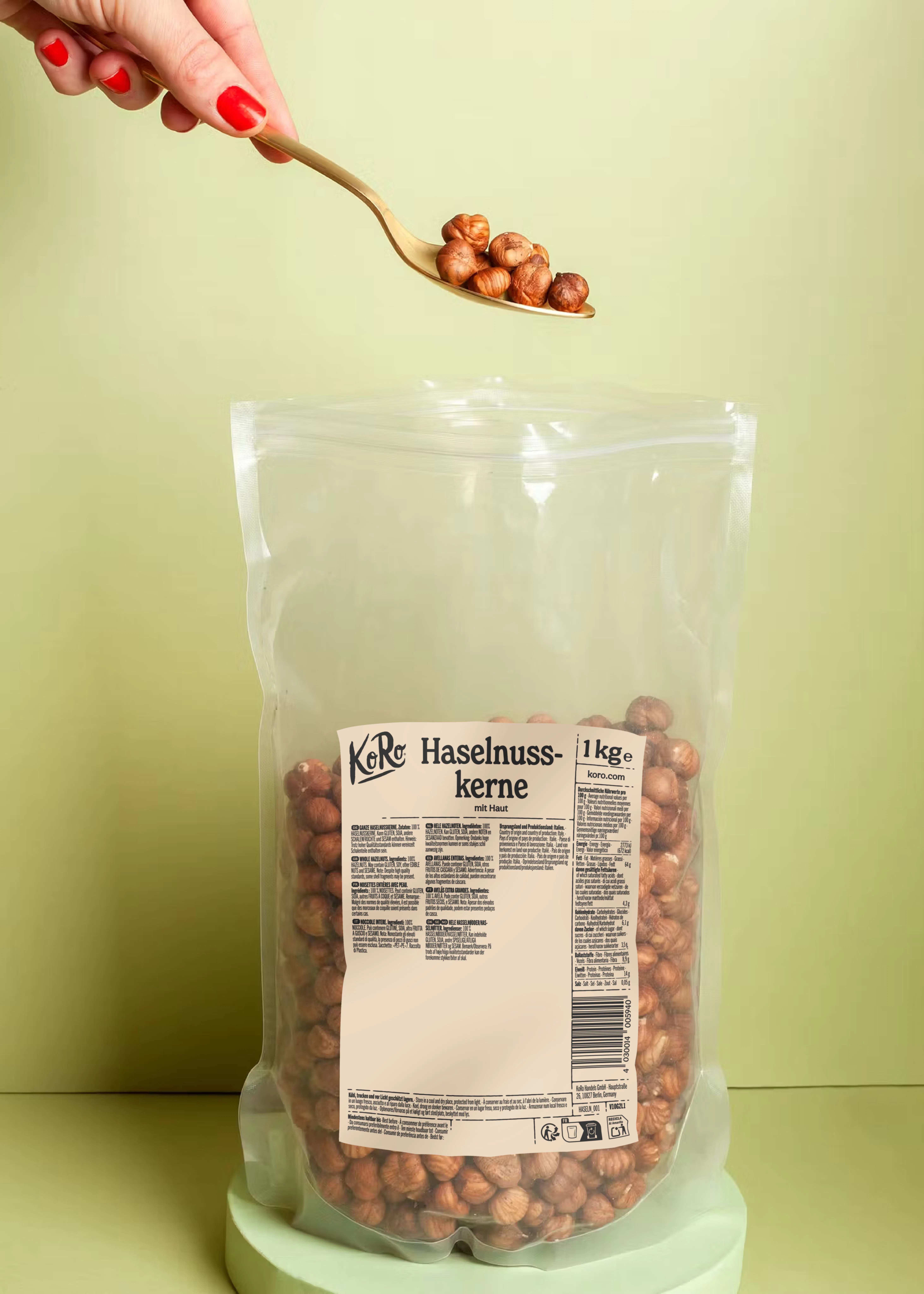 une main avec du vernis à ongles rouge tient une cuillère dorée remplie de noisettes au-dessus d'un grand sac transparent de noisettes sur un fond vert.