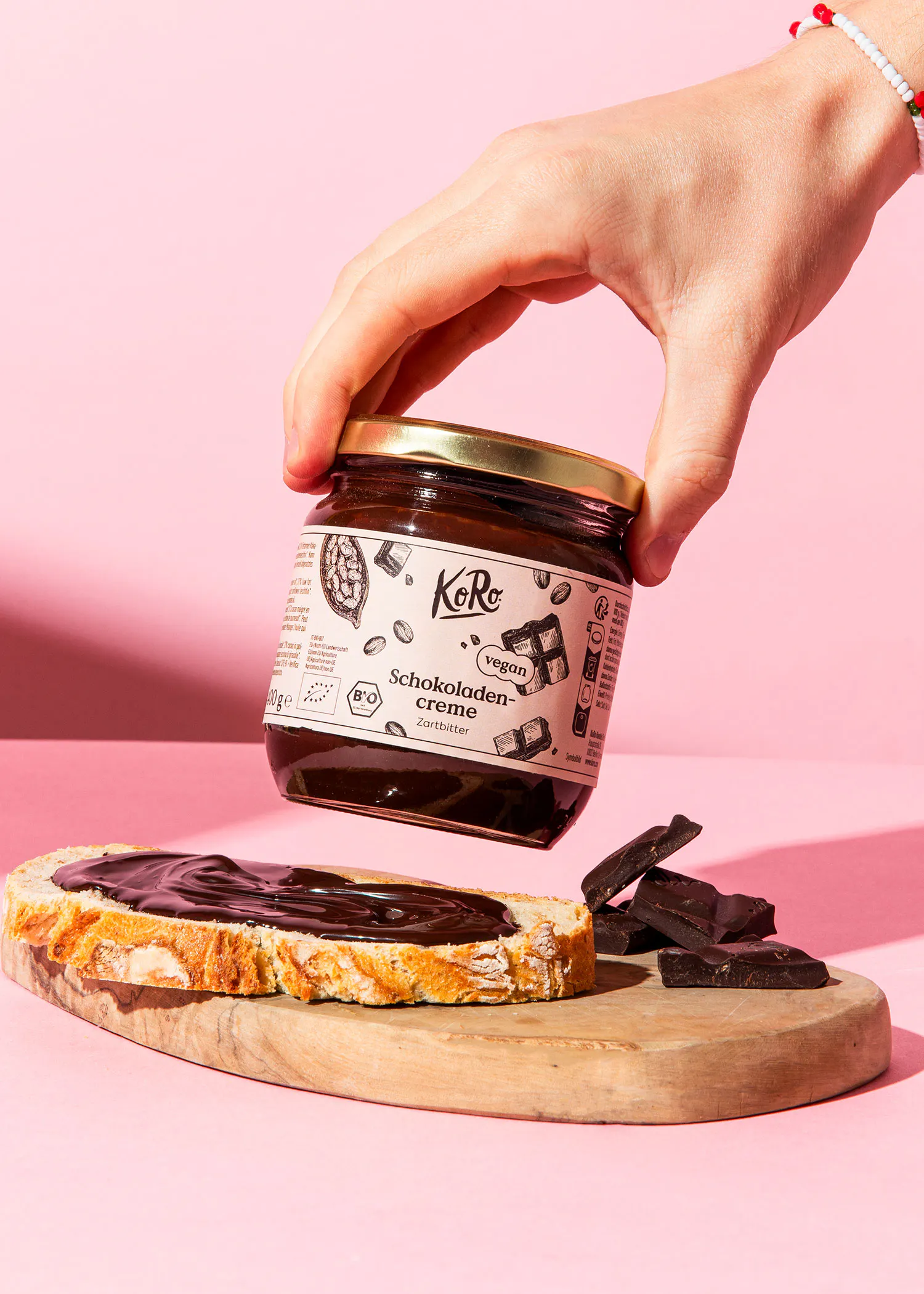 eine hand hält ein glas koro schokoladencreme zartbitter über einem brot mit schokoladencreme und schokoladenstücken auf einem holzbrett vor rosa hintergrund.