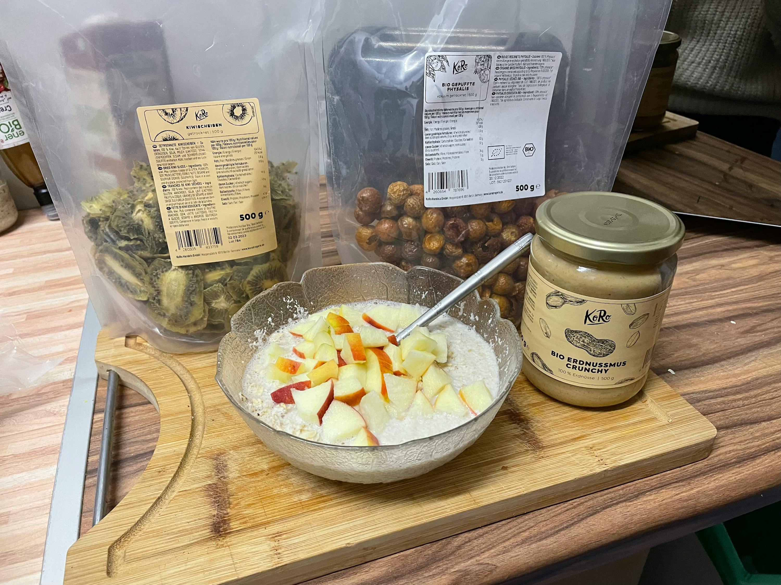 eine Schüssel Haferflocken mit geschnittenen Äpfeln steht auf einem Schneidebrett neben einem Glas bio Erdnussmus crunchy
