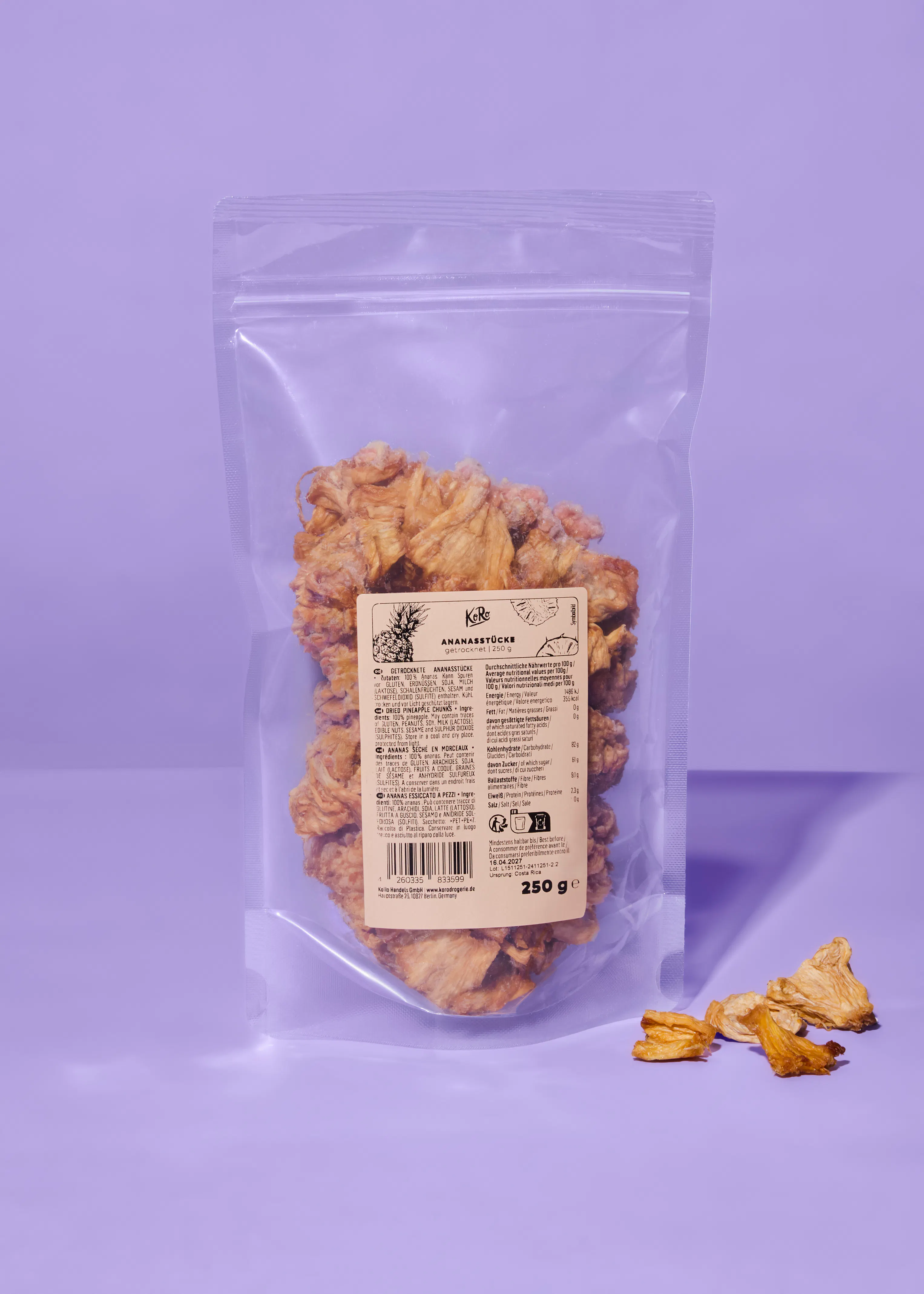 a clear plastic bag of dried pineapple chunks with a product label, and a few loose chunks, on a purple background.