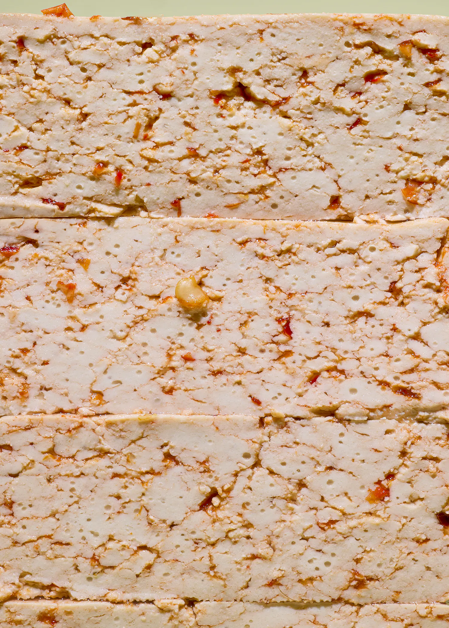 a close-up of stacked, light-colored, textured blocks of food, possibly tofu, containing small holes, red and orange flecks, and a single light-colored nut.