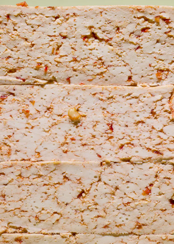 a close-up of stacked, light-colored, textured blocks of food, possibly tofu, containing small holes, red and orange flecks, and a single light-colored nut.