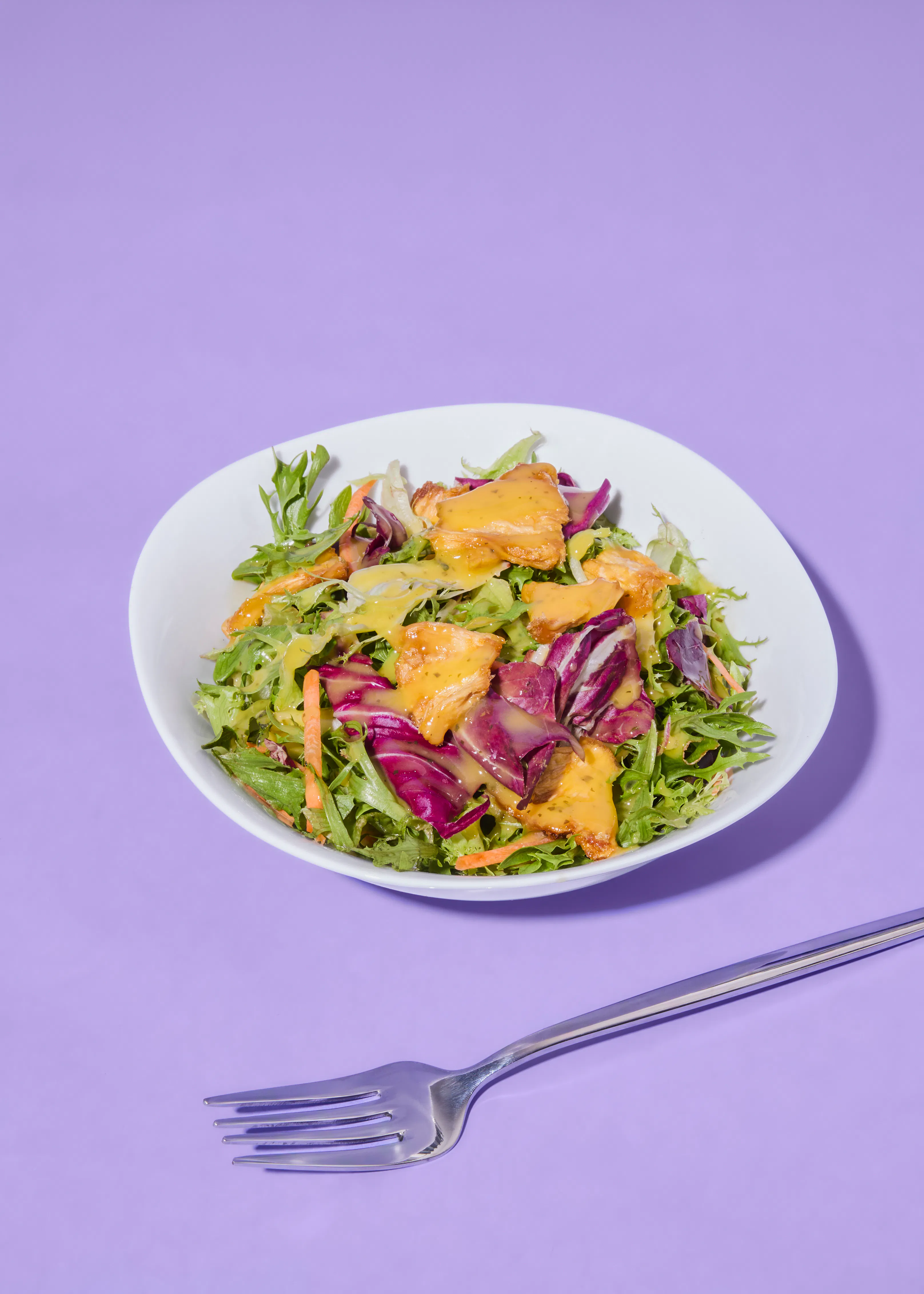 a white bowl of salad with green lettuce, red cabbage, carrots, and chicken pieces drizzled with yellow dressing, next to a silver fork on a purple background.