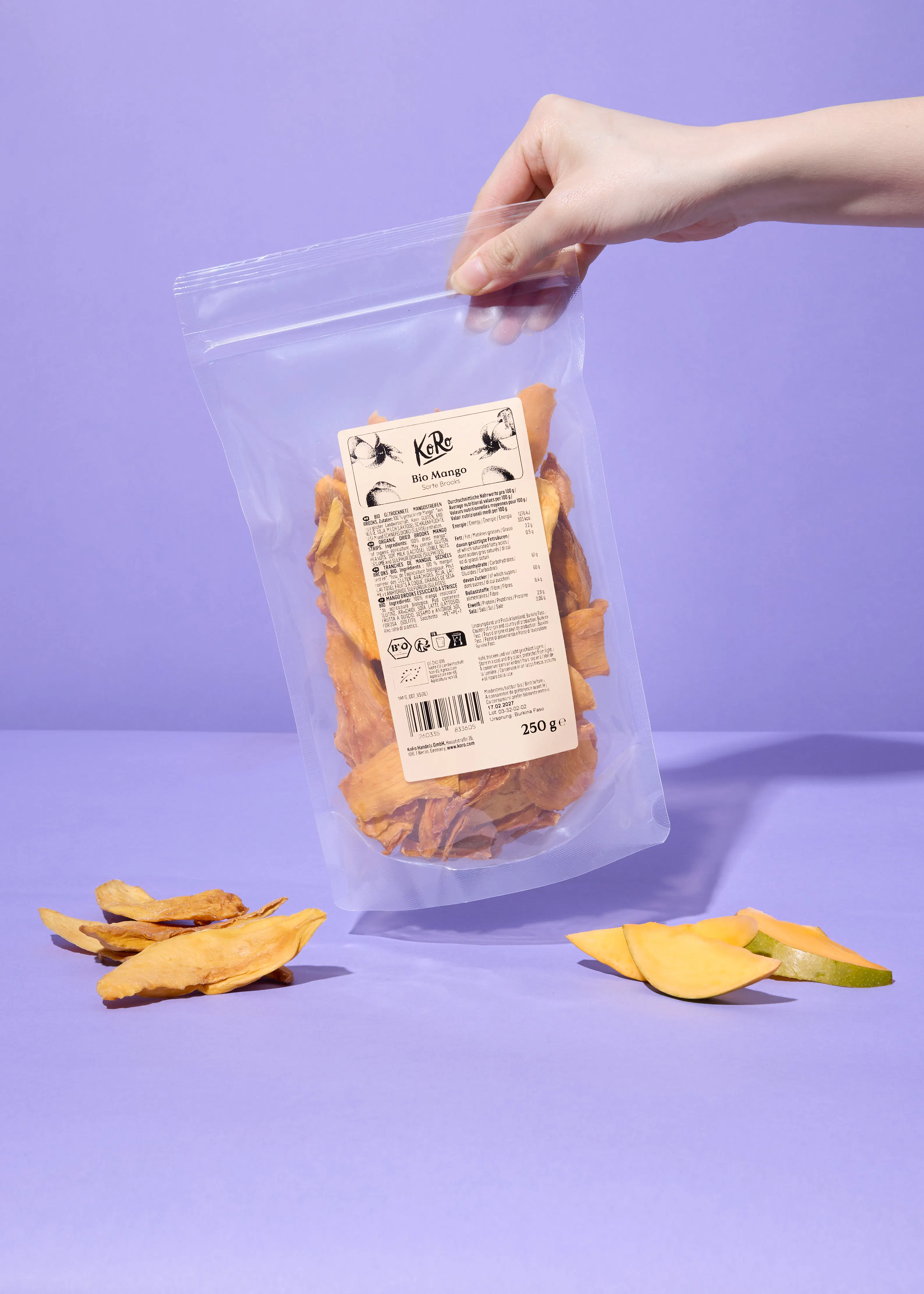 a hand holds a clear bag of koro bio mango dried slices, with fresh mango slices and more dried mango on a purple surface.