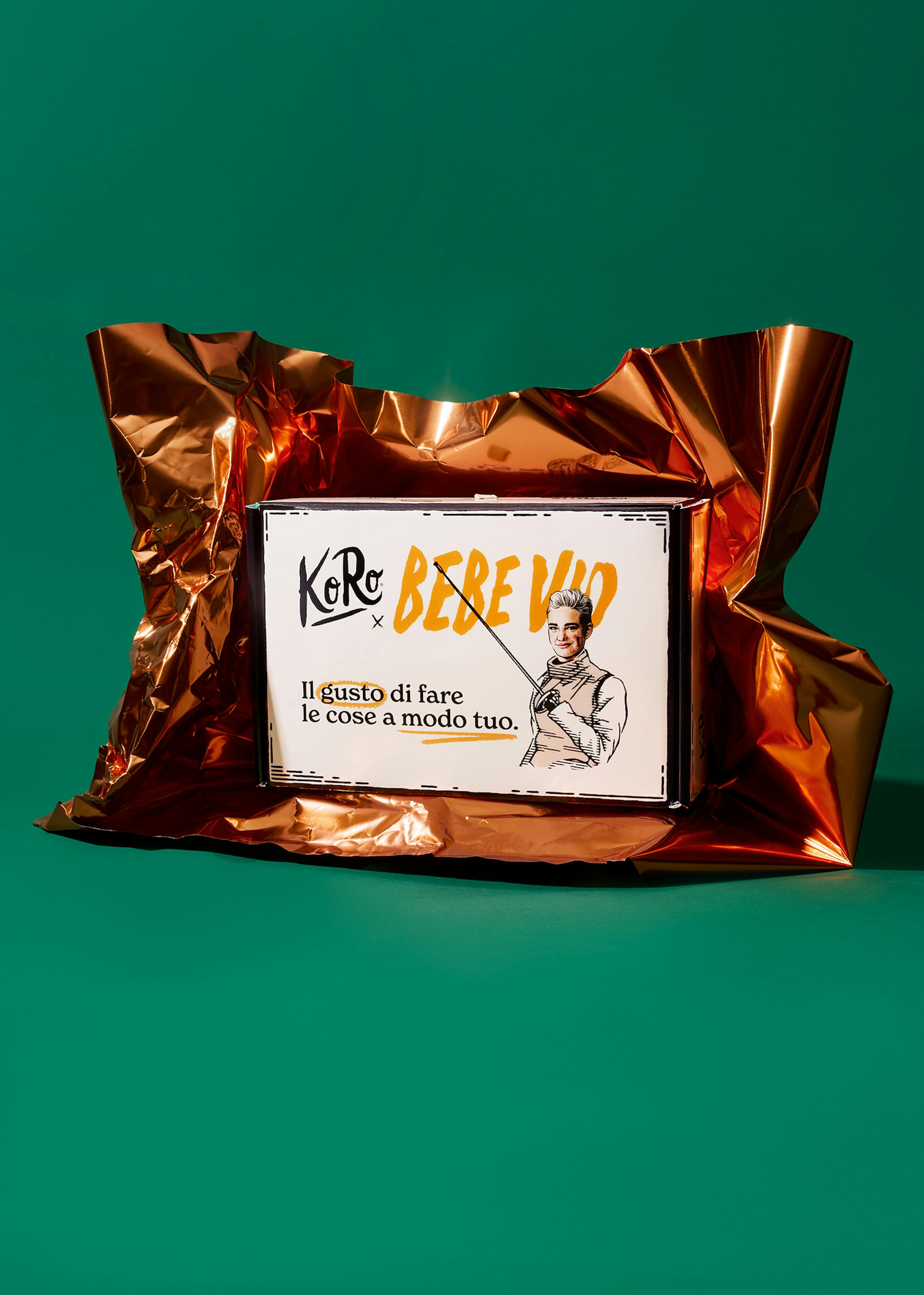 una scatola bianca avvolta in una pellicola di rame lucida, con il logo koro x bere vio, l'illustrazione di una persona che punta e la frase 'il gusto di fare le cose a modo tuo', su uno sfondo verde scuro.