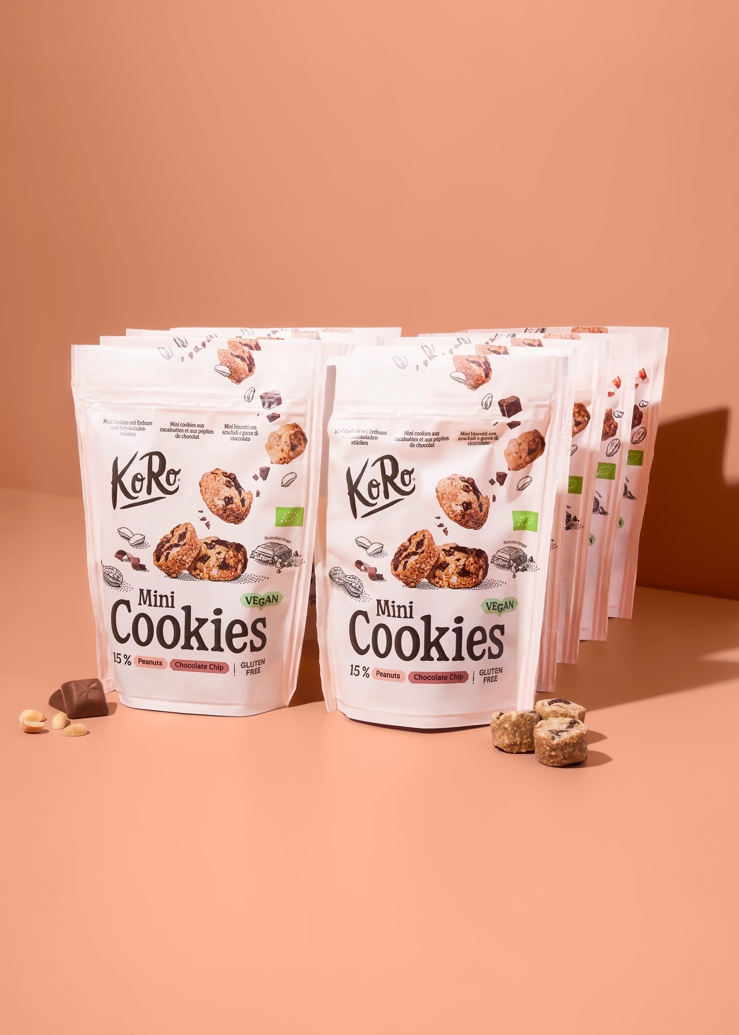 mehrere weiße standbeutel von koro mini cookies mit erdnüssen und schokoladenstückchen, die vegan und glutenfrei sind, stehen auf einer hellbraunen oberfläche, mit einigen losen erdnüssen, schokoladenstücken und keksen im vordergrund.