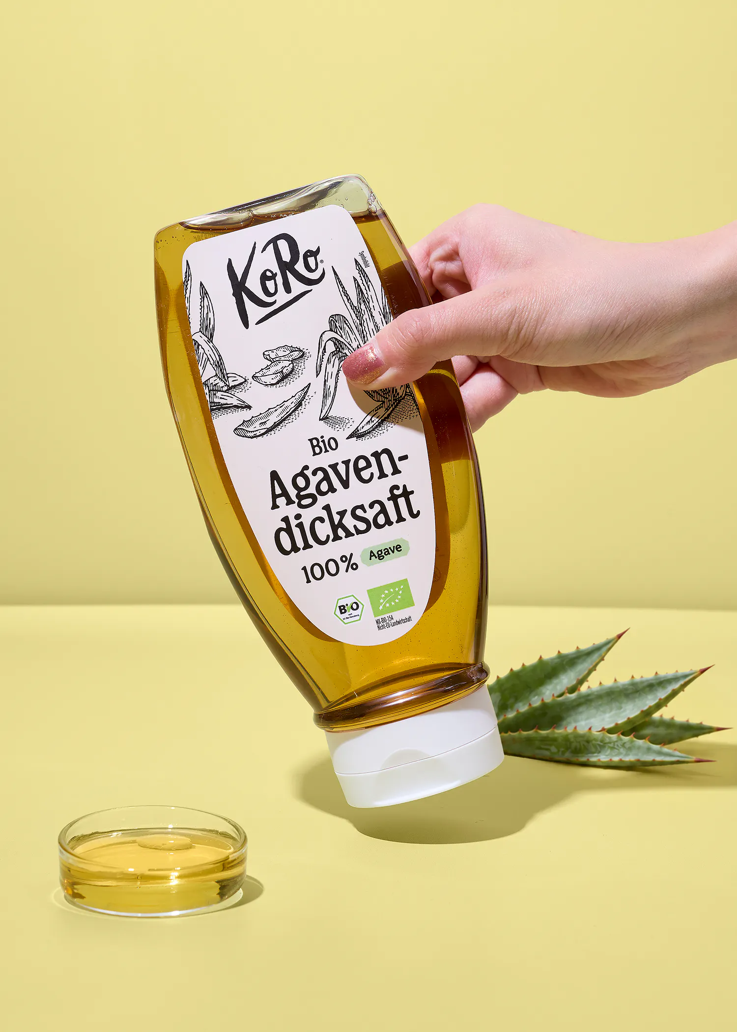 une main tient une bouteille de sirop d'agave bio koro au-dessus d'un petit récipient rempli de sirop avec des feuilles d'agave en arrière-plan sur un fond jaune.