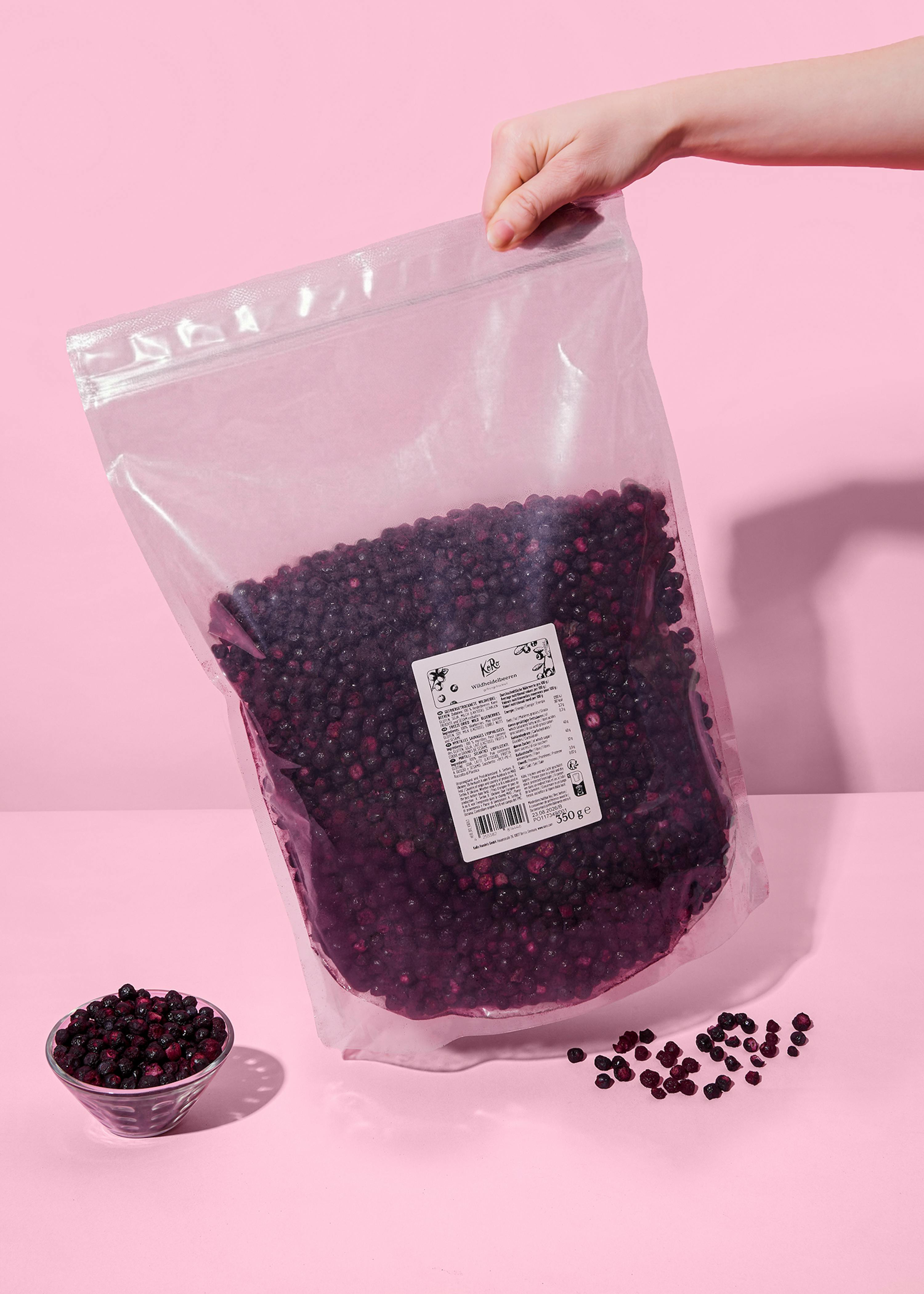 a hand holds a large clear bag of frozen wild blueberries, with a small bowl and scattered blueberries on a pink background.
