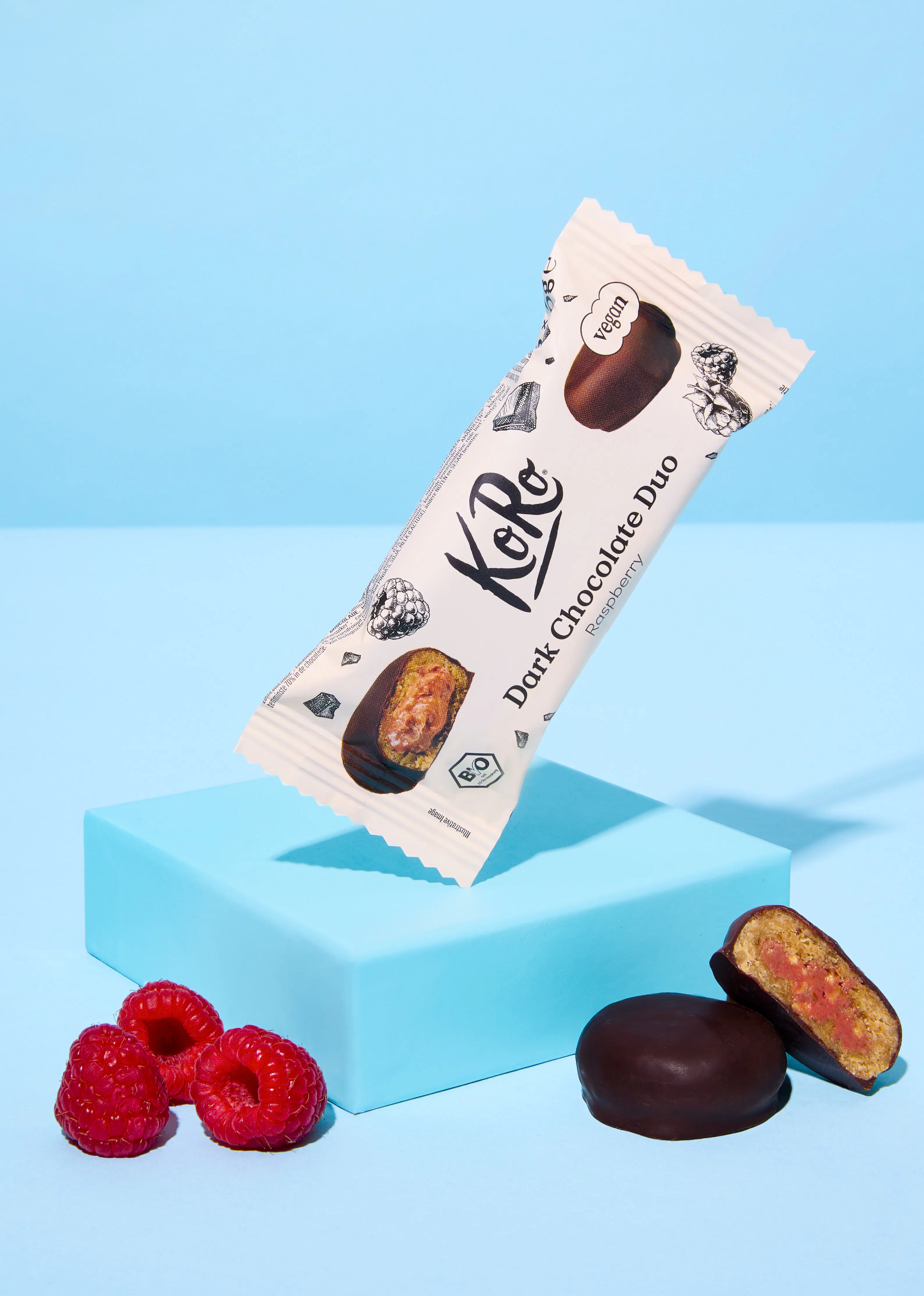 eine teilweise ausgepackte koro dark chocolate duo himbeer-riegelpackung steht auf einem hellblauen block, daneben liegen frische himbeeren und zwei schokoladenstücke, eines davon aufgeschnitten mit rosa füllung.