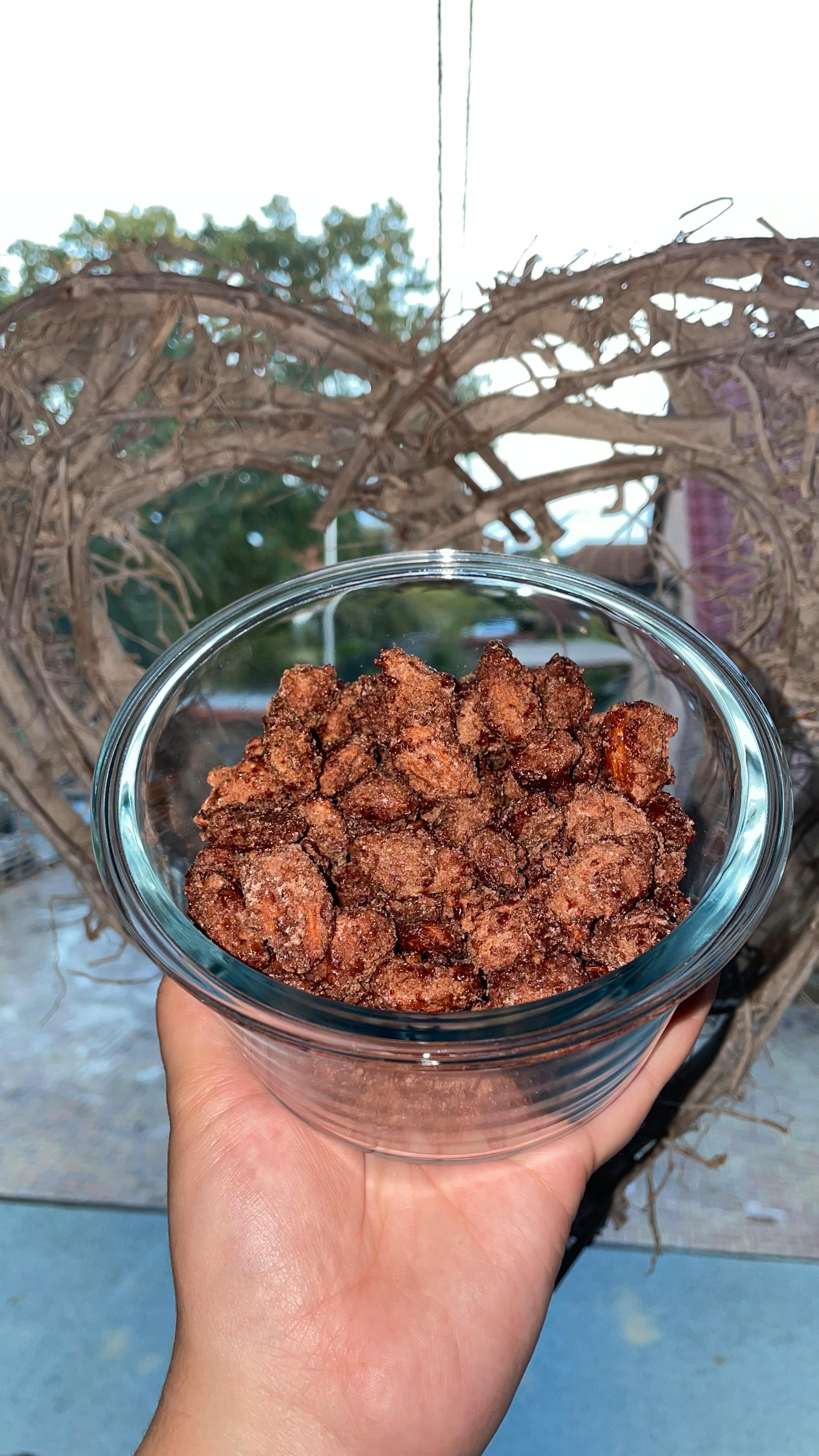 a hand holds a clear glass bowl filled with candied nuts, with a rustic heart-shaped wreath in the background.