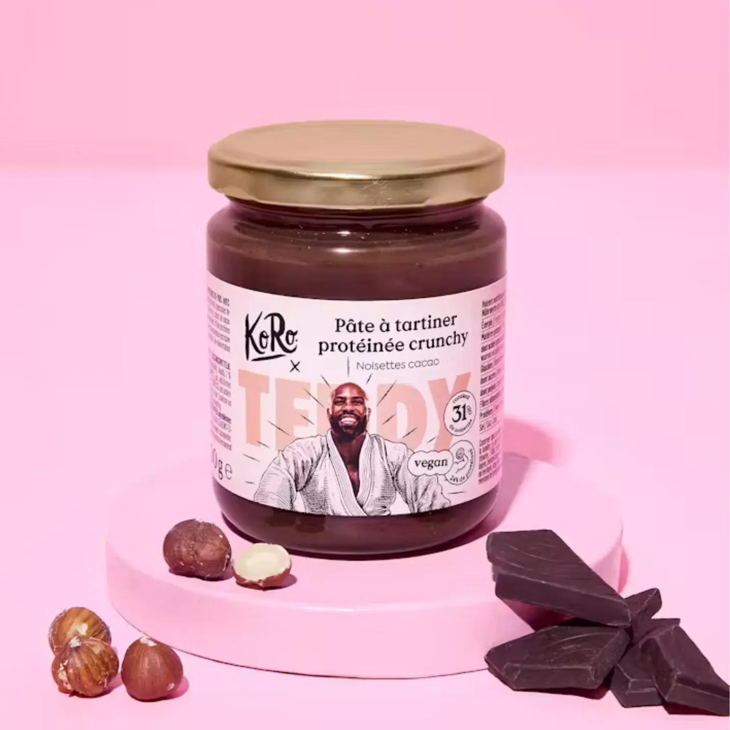 pot de pâte à tartiner protéinée crunchy koro aux noisettes et au cacao avec une illustration de teddy riner sur l'étiquette, posé sur un socle rose entouré de noisettes et de morceaux de chocolat noir.