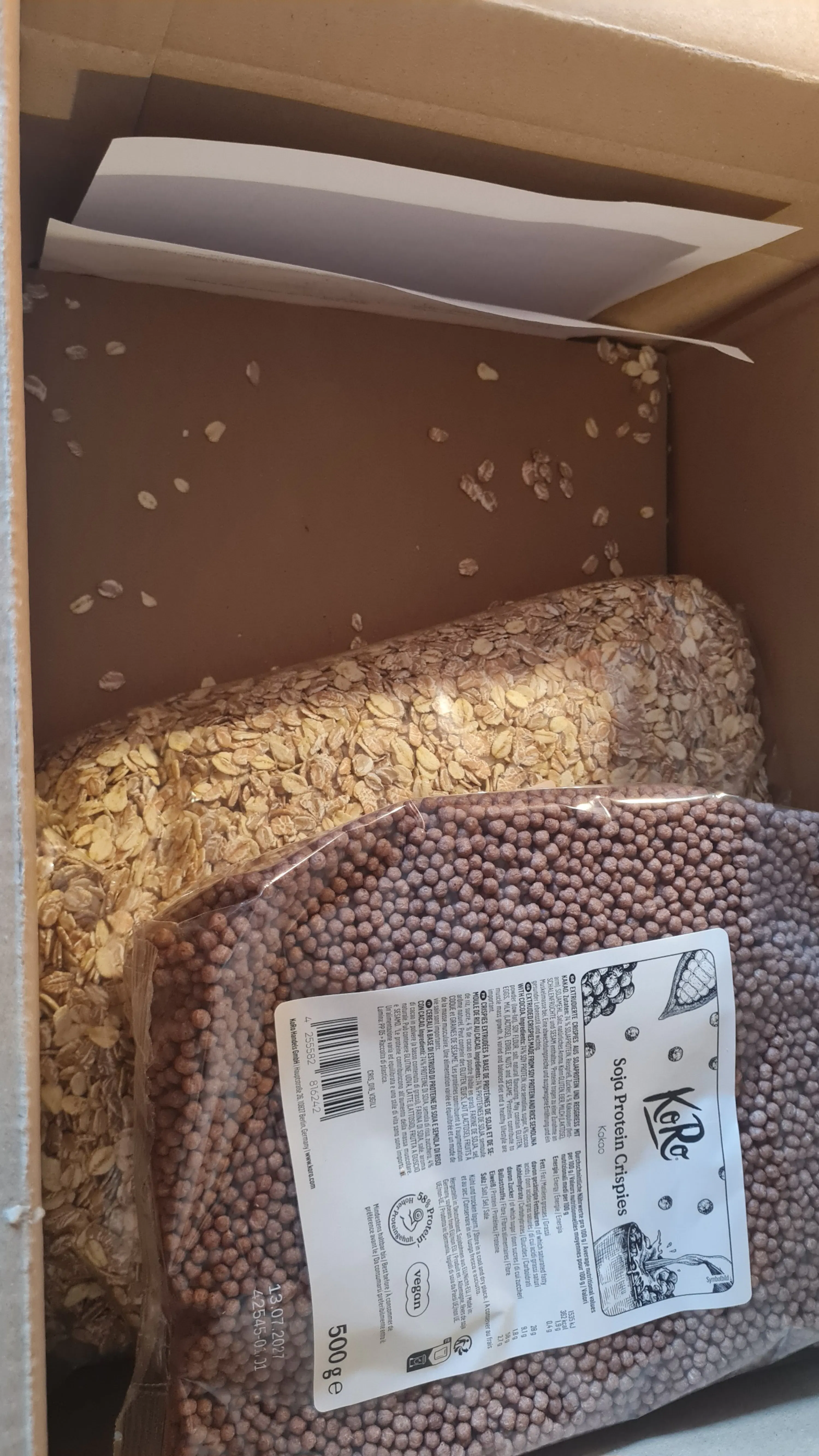 eine offene braune versandbox, die zwei durchsichtige beutel mit lebensmitteln enthält: einen beutel mit hellbraunen haferflocken und einen beutel mit dunklen koro soja protein crispies kakao, mit einigen verstreuten haferflocken am boden.