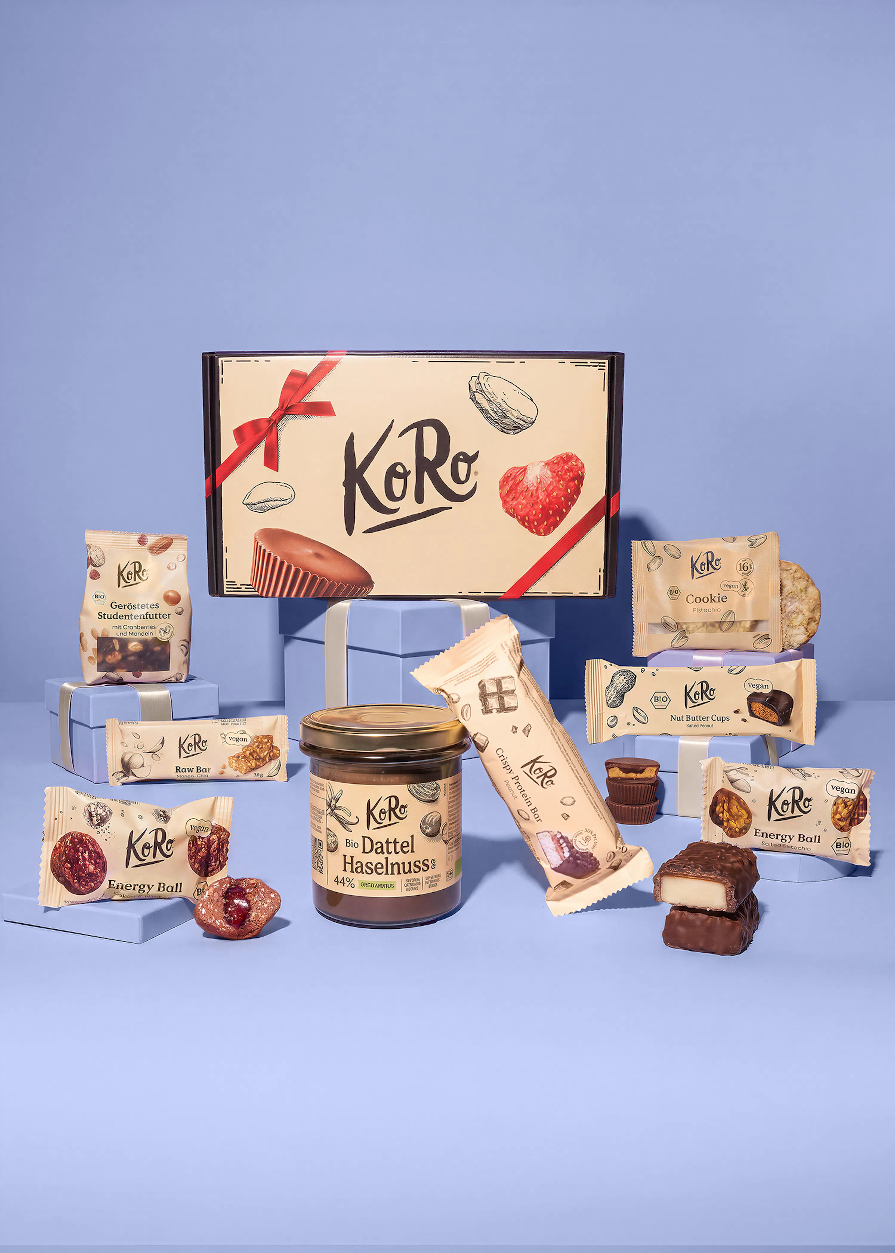 eine auswahl an koro-snackprodukten, darunter eine geschenkbox, dattel-haselnuss-creme, proteinriegel, energiekugeln und nussbutter-cups, präsentiert auf hellblauen podesten vor blauem hintergrund.
