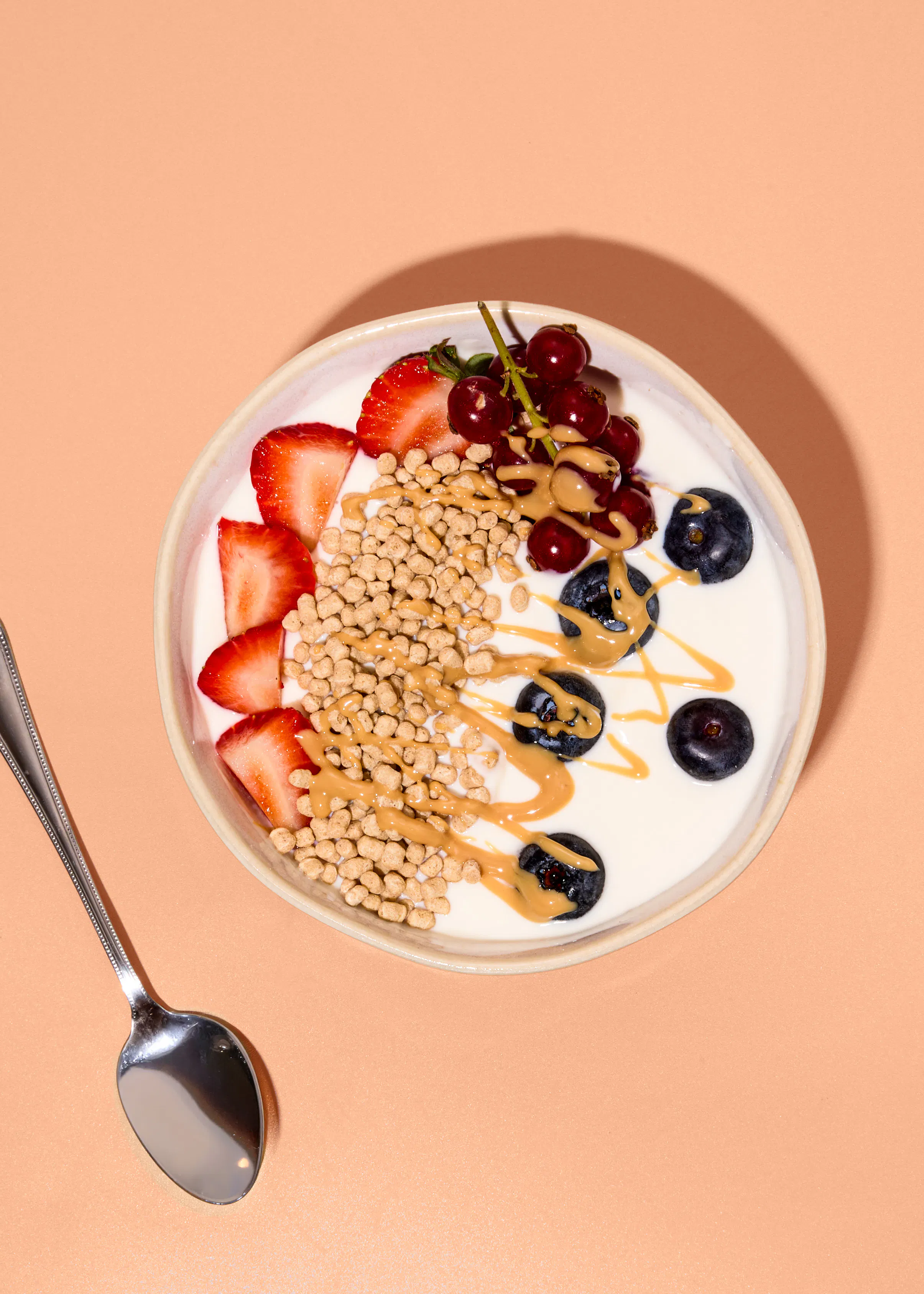 eine schüssel mit joghurt, garniert mit erdbeerscheiben, heidelbeeren, johannisbeeren, knuspermüsli und einem drizzle aus erdnussbutter, daneben liegt ein silberner löffel auf pfirsichfarbenem hintergrund.