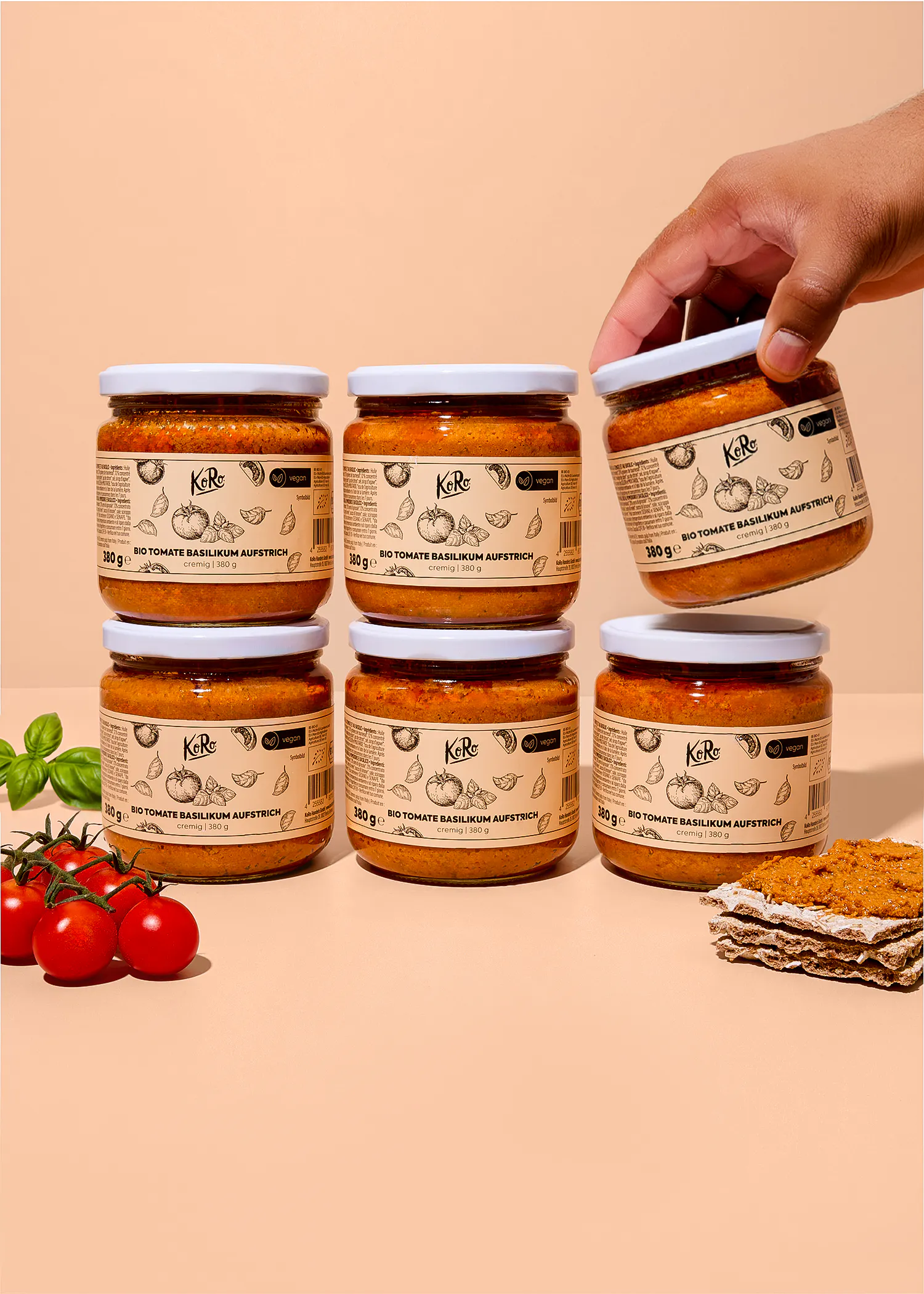 una mano solleva un barattolo di crema spalmabile koro al pomodoro e basilico sopra una pila di altri cinque barattoli identici, con pomodorini freschi e una fetta di pane croccante accanto.