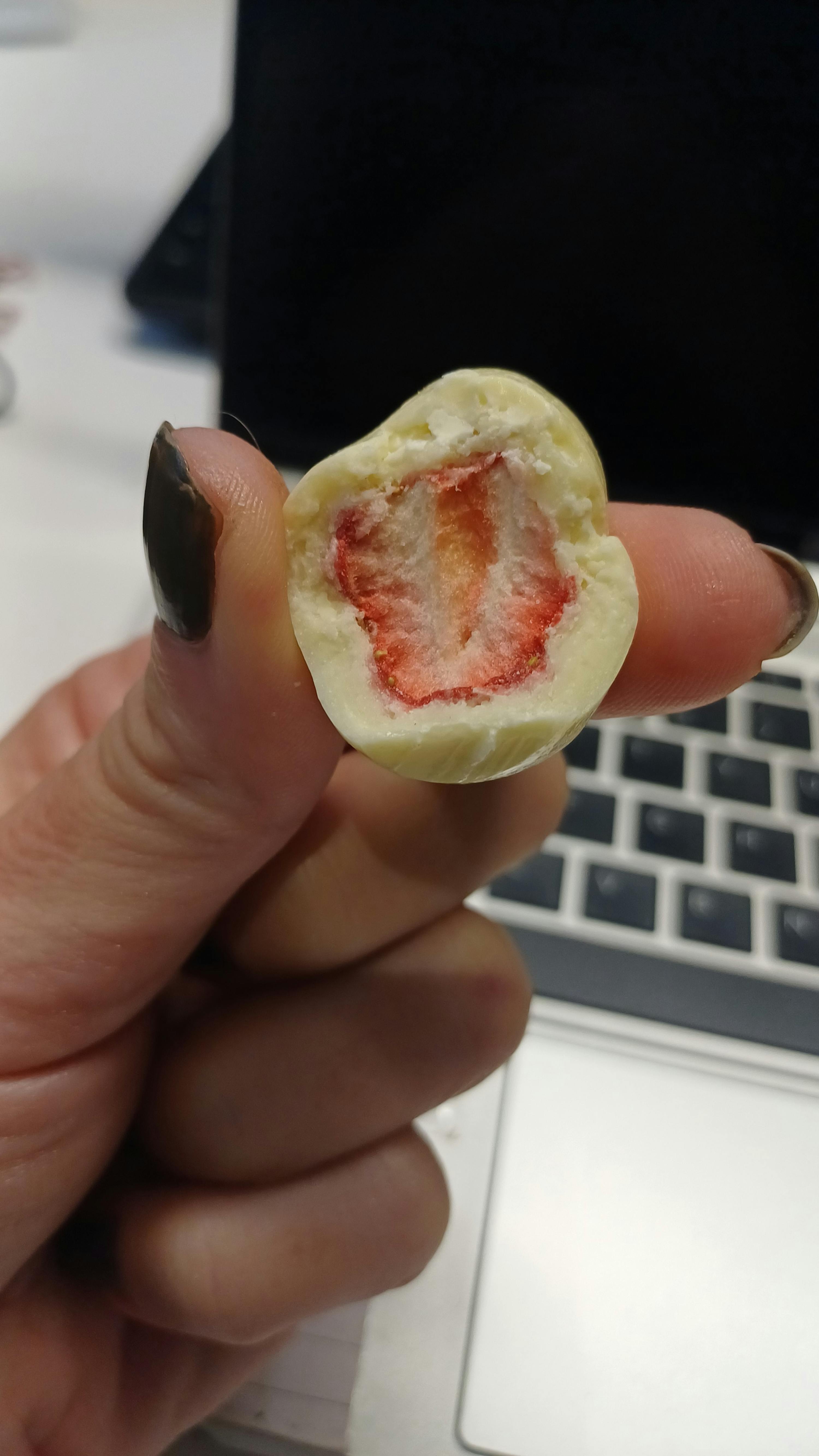 une main tient un bonbon au chocolat blanc coupé en deux, révélant un centre de fraise lyophilisée, avec un clavier d'ordinateur flou en arrière-plan.