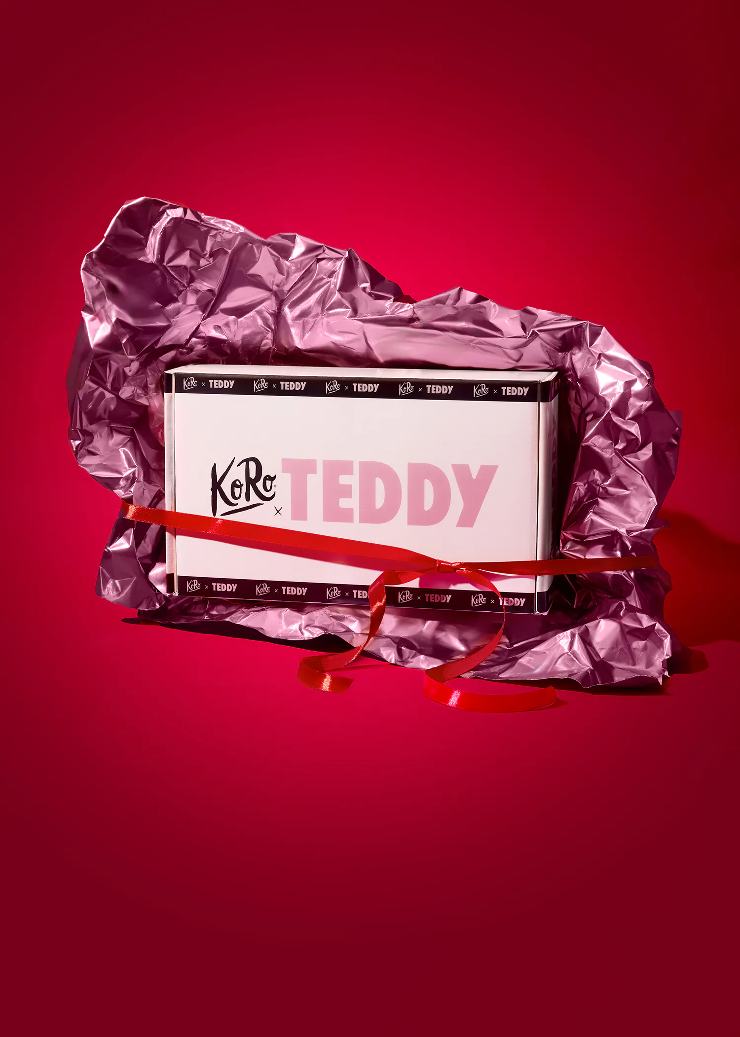 une boîte blanche de la marque koro x teddy, emballée dans du papier métallique rose froissé et nouée avec un ruban rouge, sur un fond rouge.