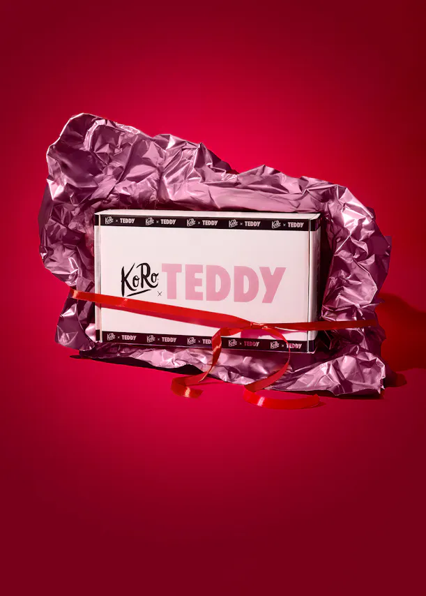 une boîte blanche de la marque koro x teddy, emballée dans du papier métallique rose froissé et nouée avec un ruban rouge, sur un fond rouge.