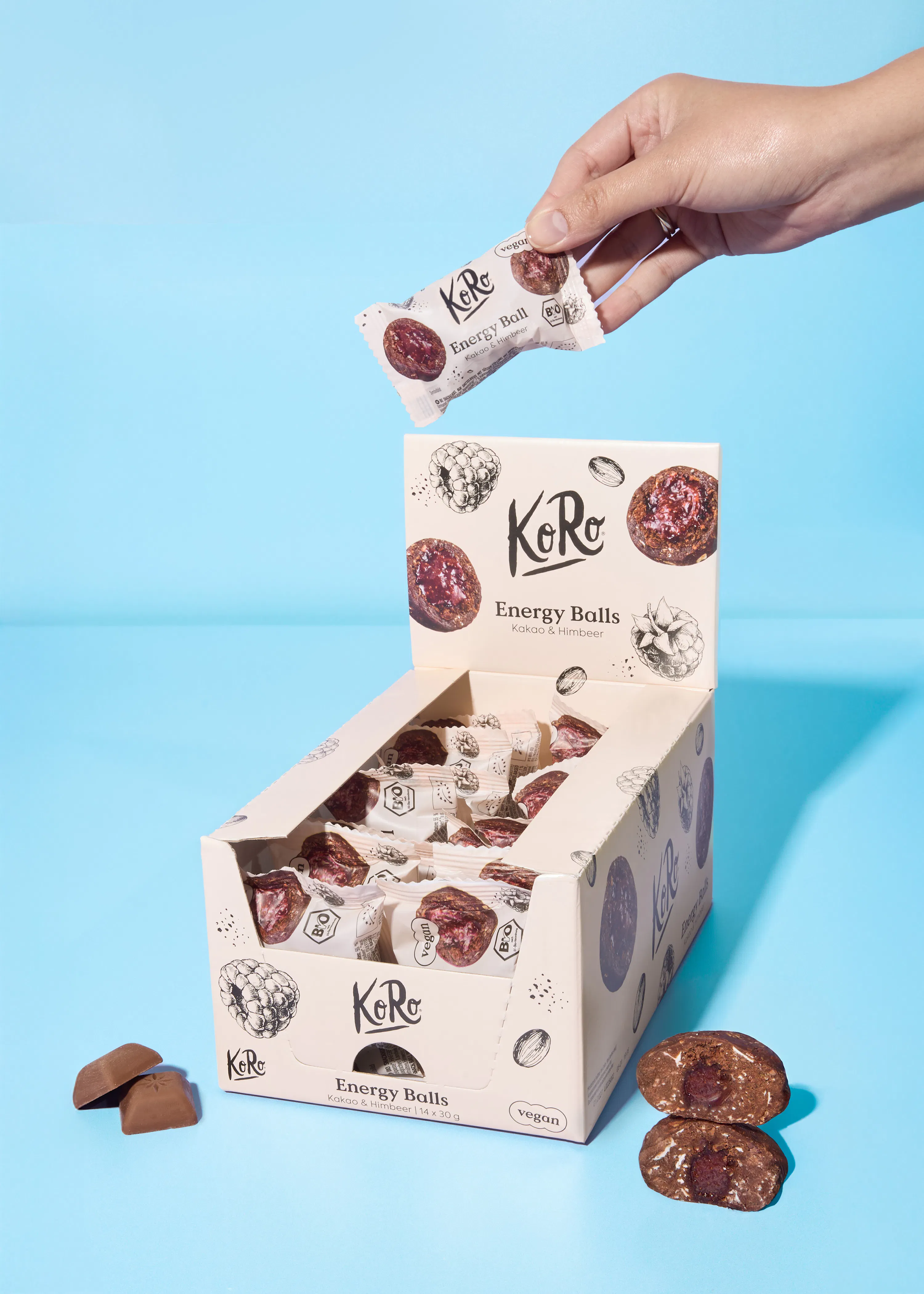 une main prend une boule d'énergie koro saveur cacao et framboise d'une boîte de présentation remplie de boules emballées, avec des boules coupées et des chocolats à côté, sur un fond bleu.