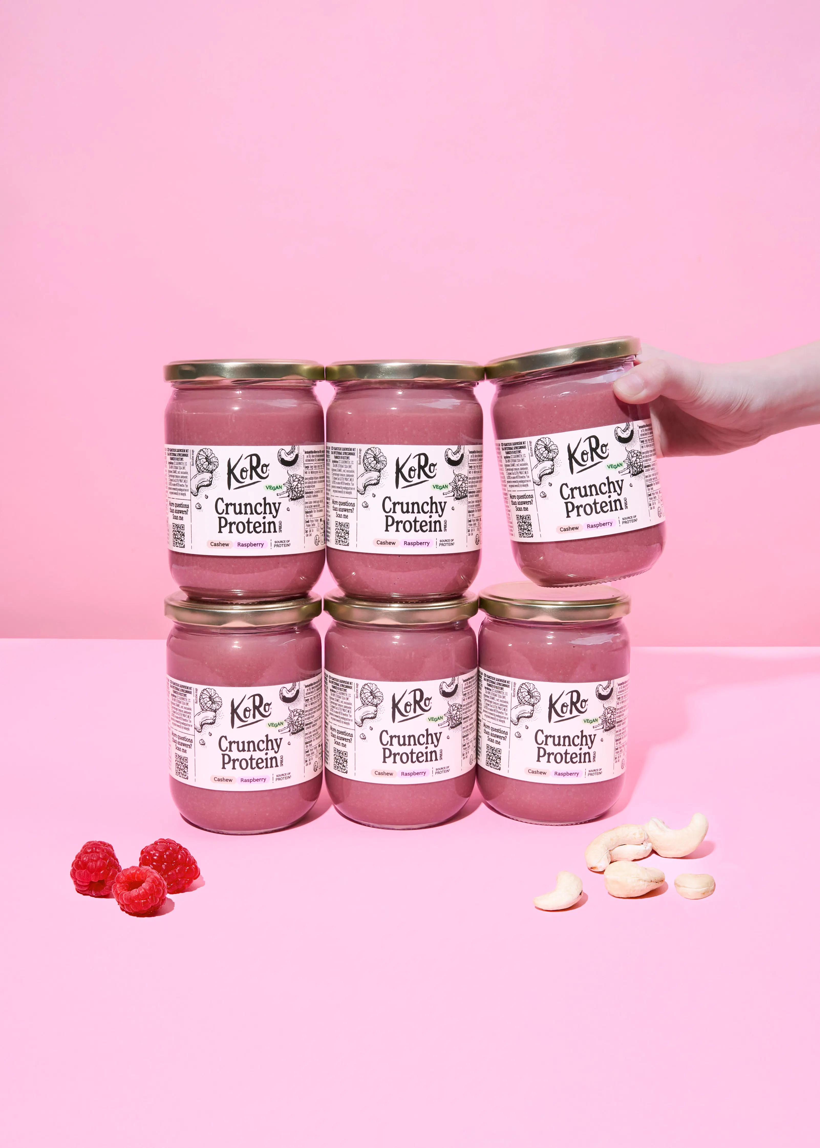 sechs gläser koro crunchy protein cashew-himbeer-aufstrich auf einem pinken hintergrund, wobei ein glas von einer hand gehalten wird und frische himbeeren sowie cashewnüsse daneben liegen.